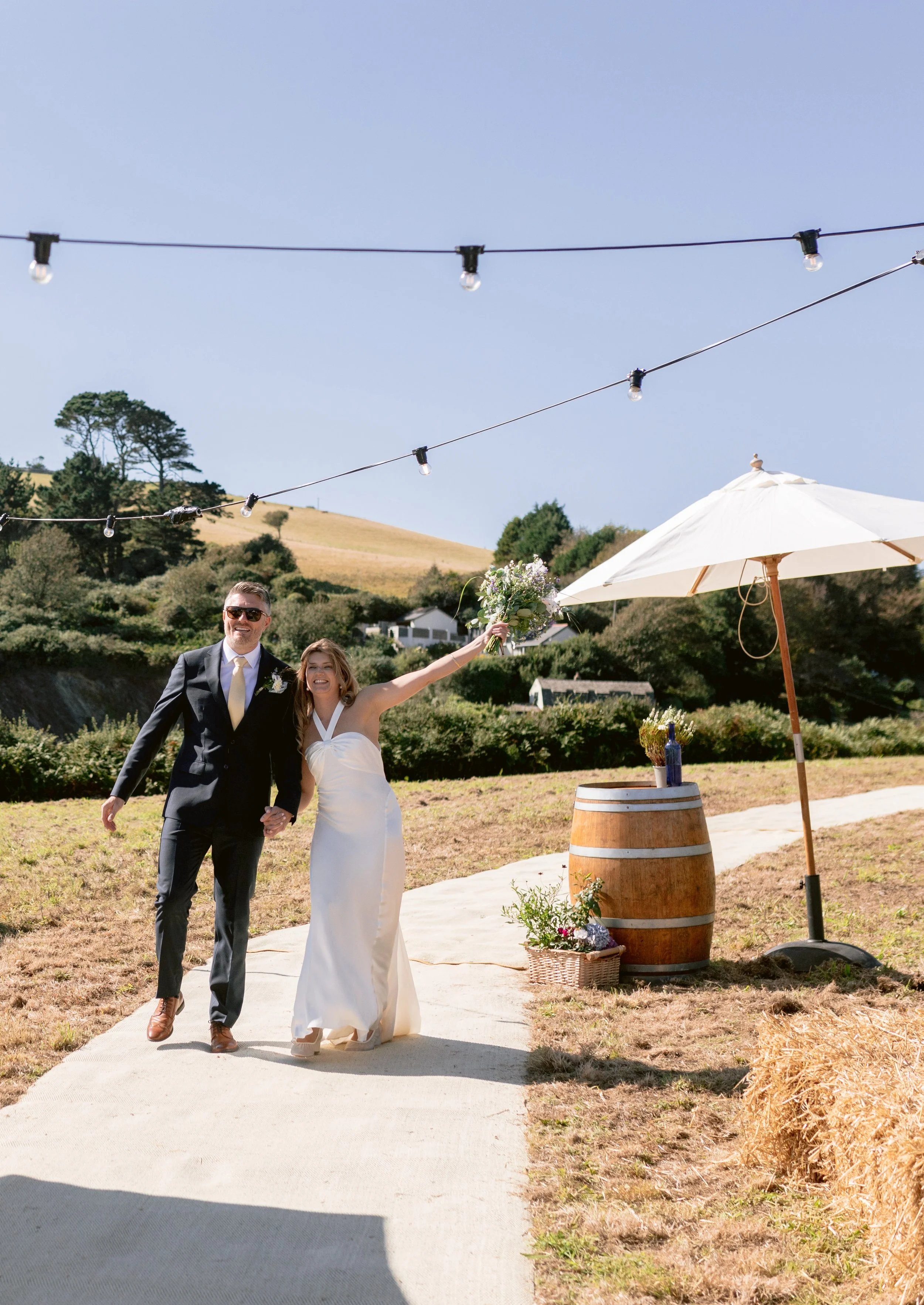A newlywed couple walking outdoors on a sunny day, with the bride holding a bouquet and gesturing happily, under string lights and near a table with a bottle, with hills and trees in the background.