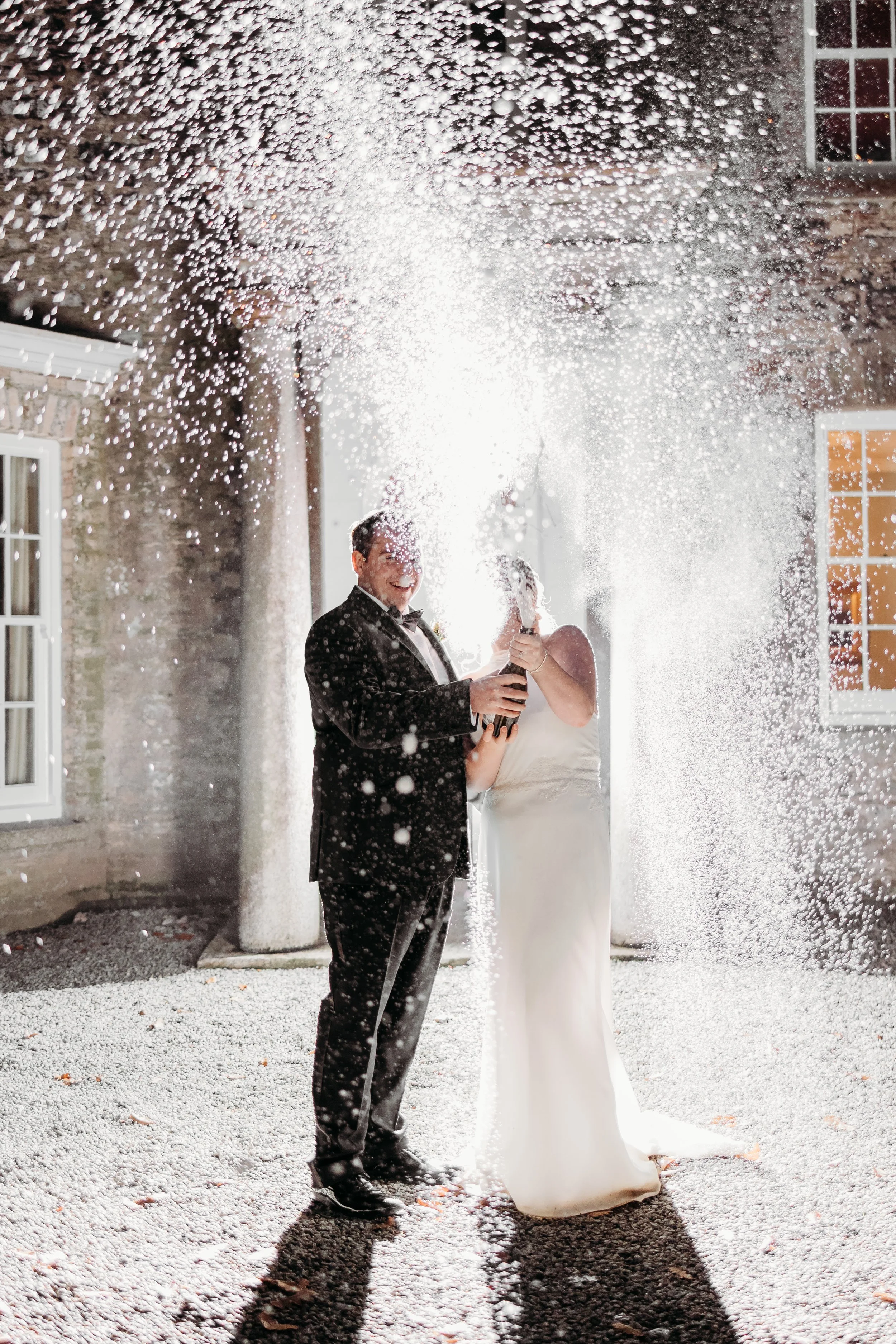 A bride and groom celebrating their wedding outdoors at night, holding a champagne bottle and spraying champagne, surrounded by falling confetti or snow.