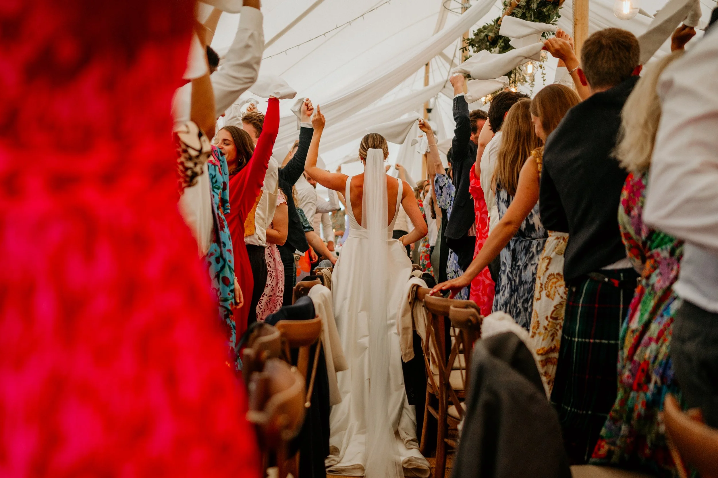 Bride in a white wedding dress walking down the aisle at her wedding reception, surrounded by guests holding white napkins or tissues, inside a decorated tent.