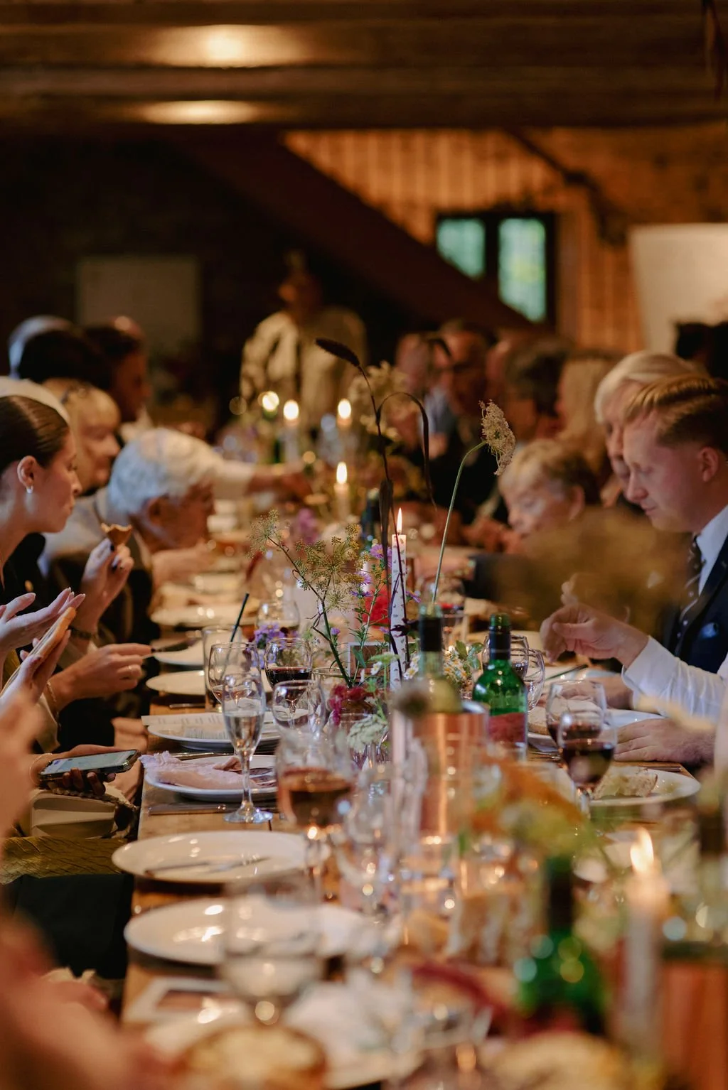 A long dining table set for a formal meal with glasses, plates, and floral centerpieces inside a rustic wooden room, with people engaged in eating and talking.