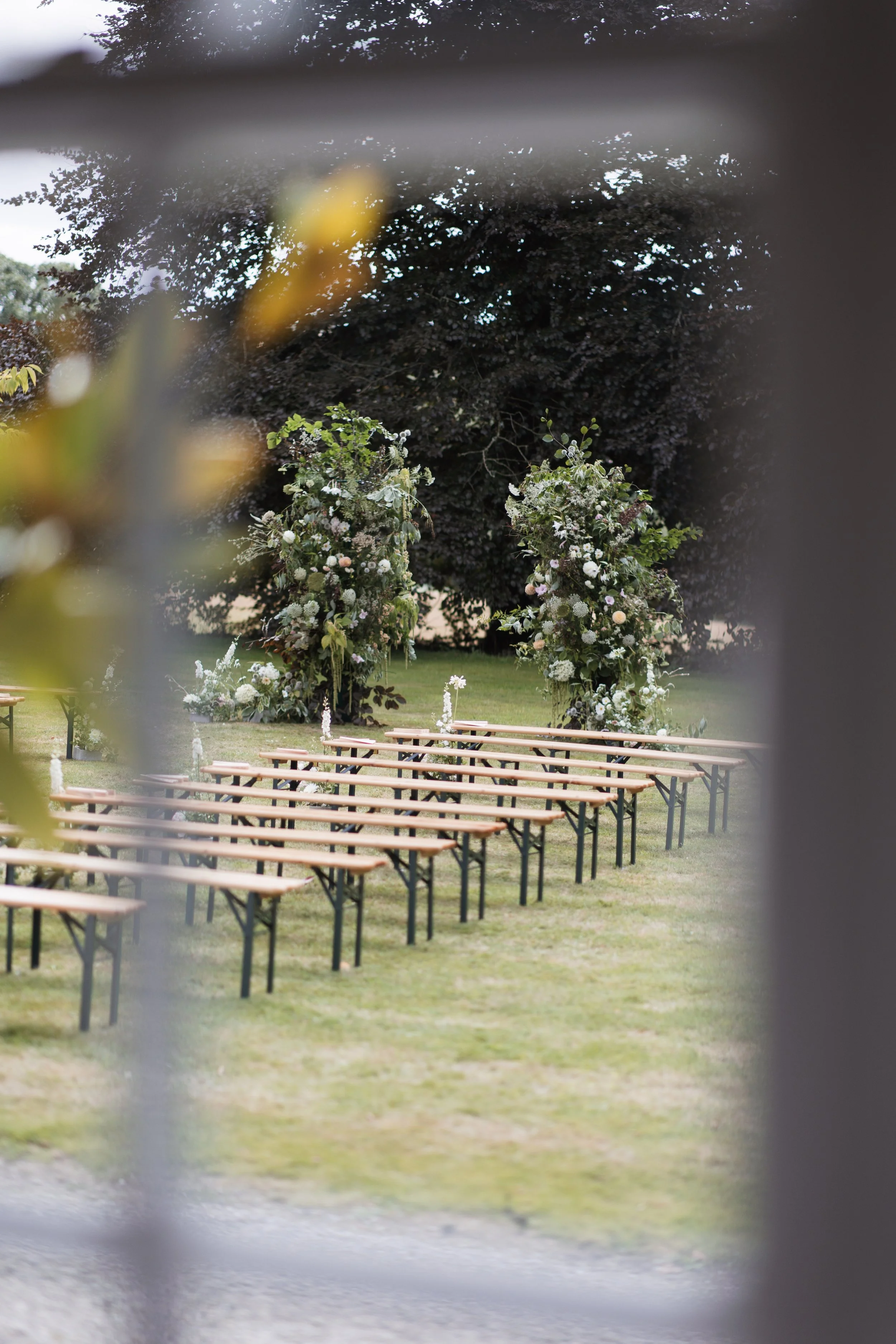 Outdoor ceremony setup with wooden benches and floral arrangements, viewed through a window.