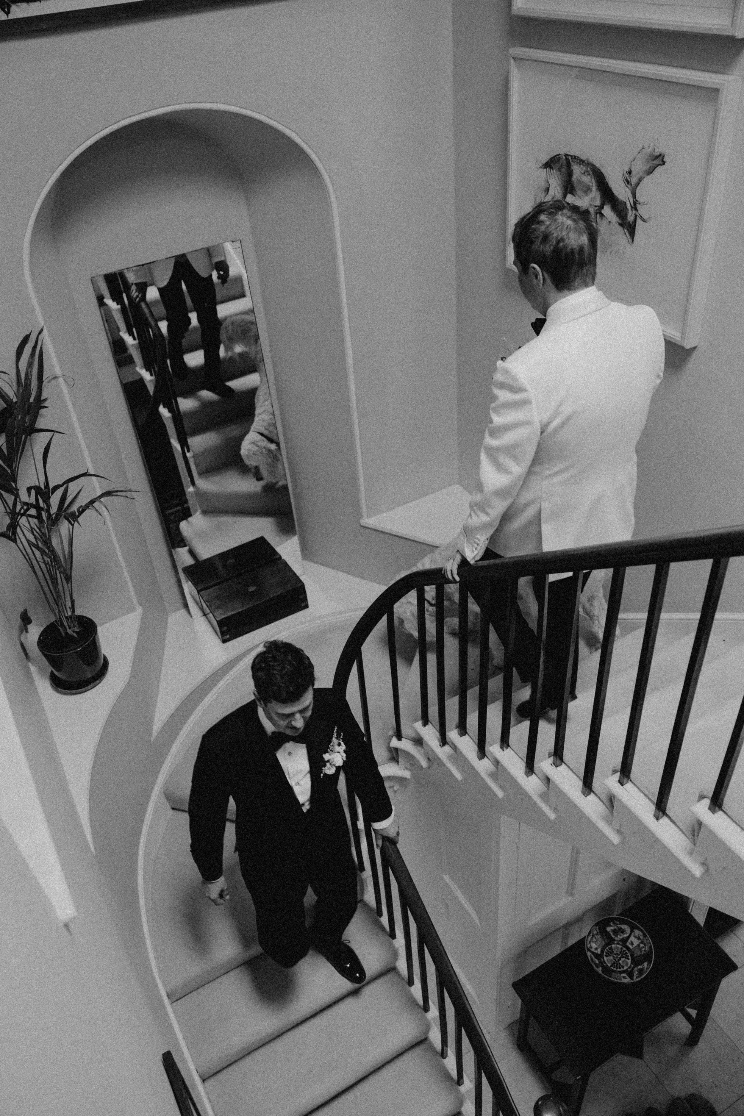 Black and white photo of two men in tuxedos descending a staircase, with one man at the top in a white tuxedo and the other below in a black tuxedo, and a woman at the top of the stairs adjusting her shoes.