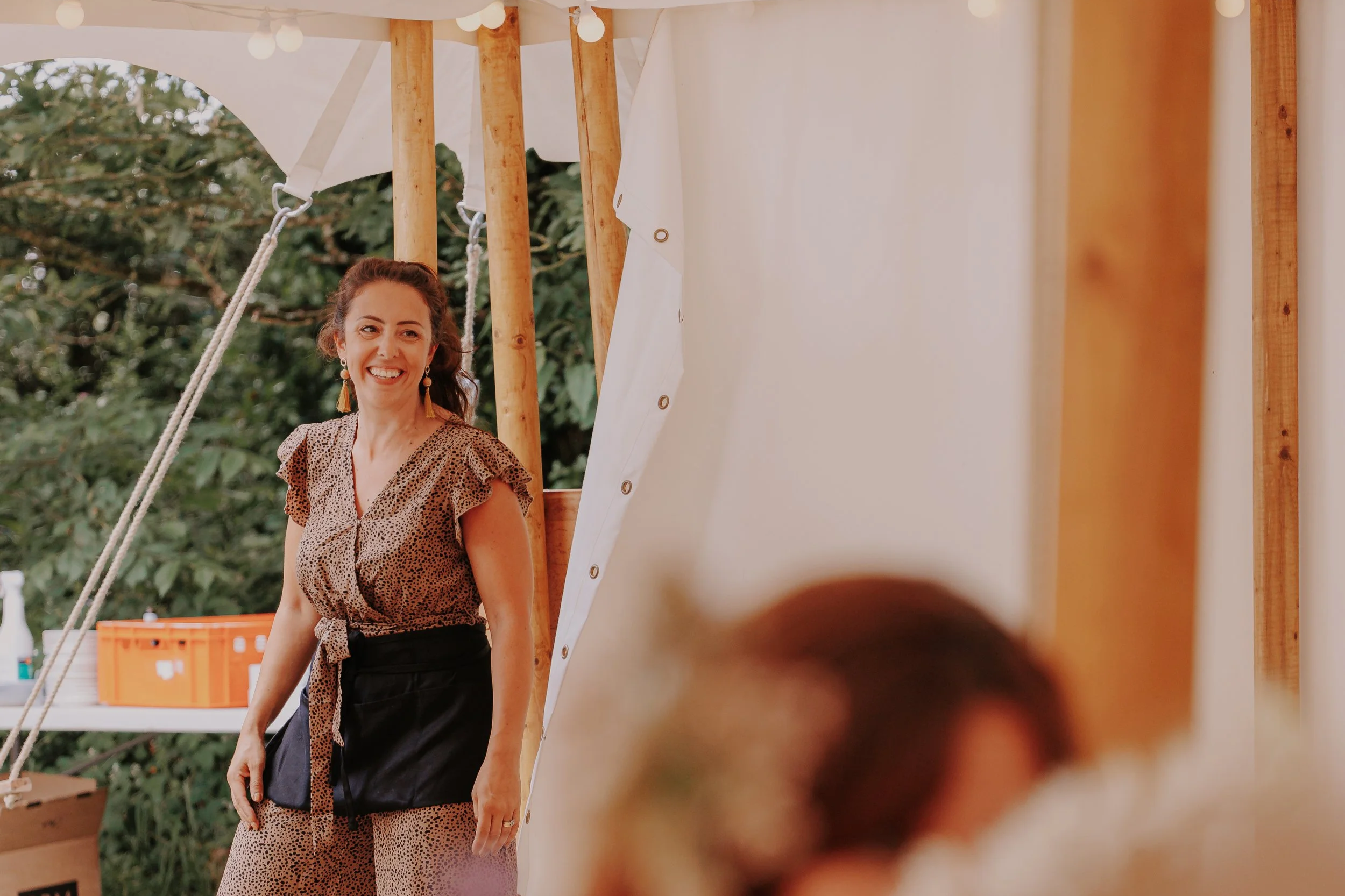 Smiling woman in a patterned dress and earrings standing outdoors near wooden structure with white fabric and greenery in background.