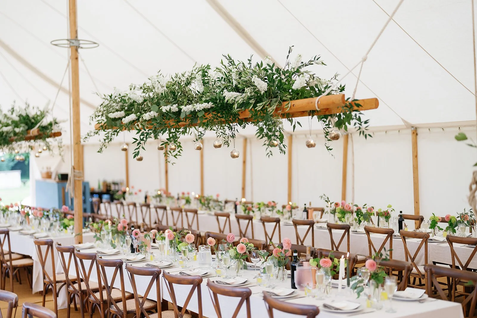 Long banquet table decorated with pink flowers and greenery under hanging floral arrangements in a large white tent.