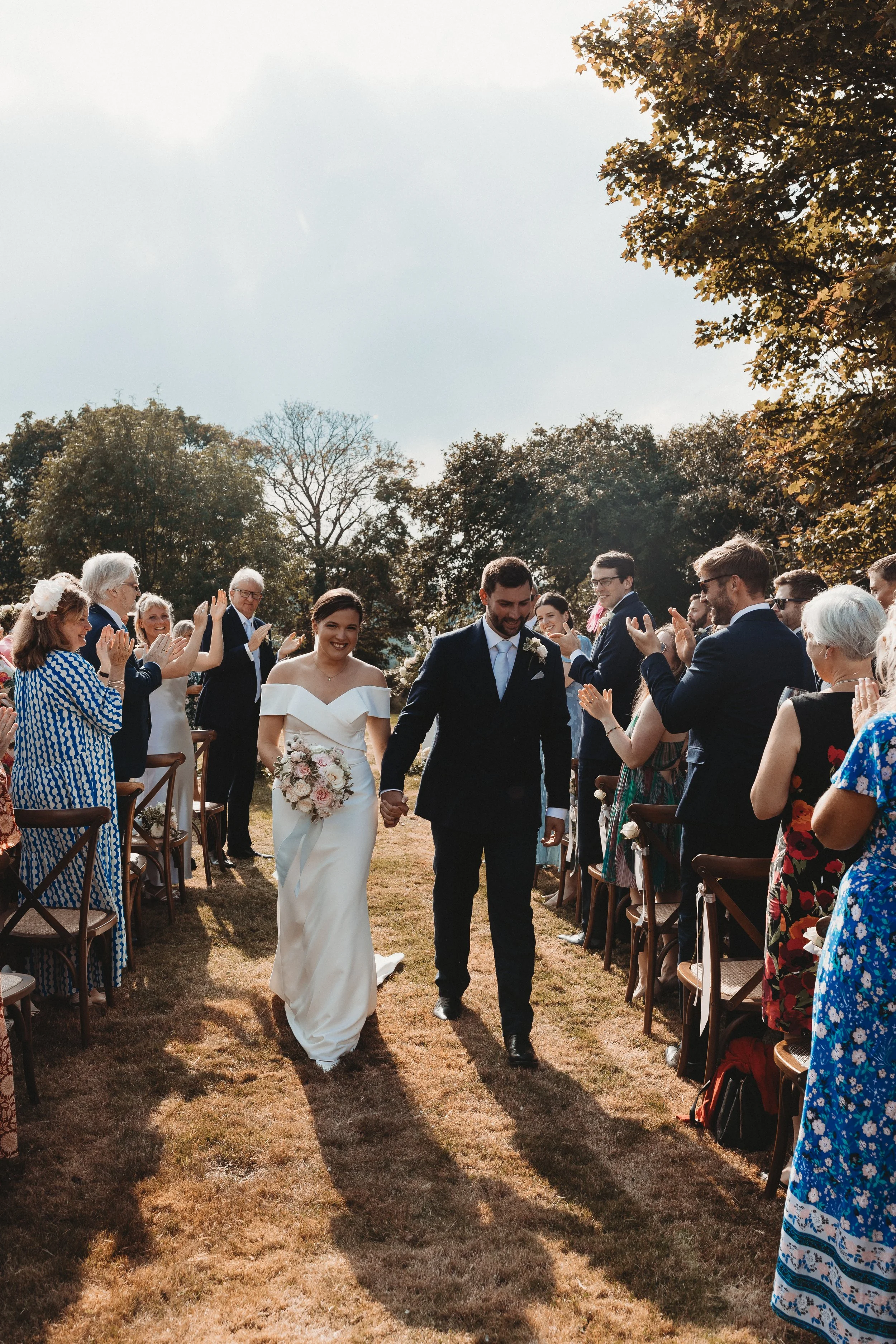 A newly married couple walking hand in hand down an outdoor aisle surrounded by friends and family, celebrating their wedding on a sunny day with trees in the background.