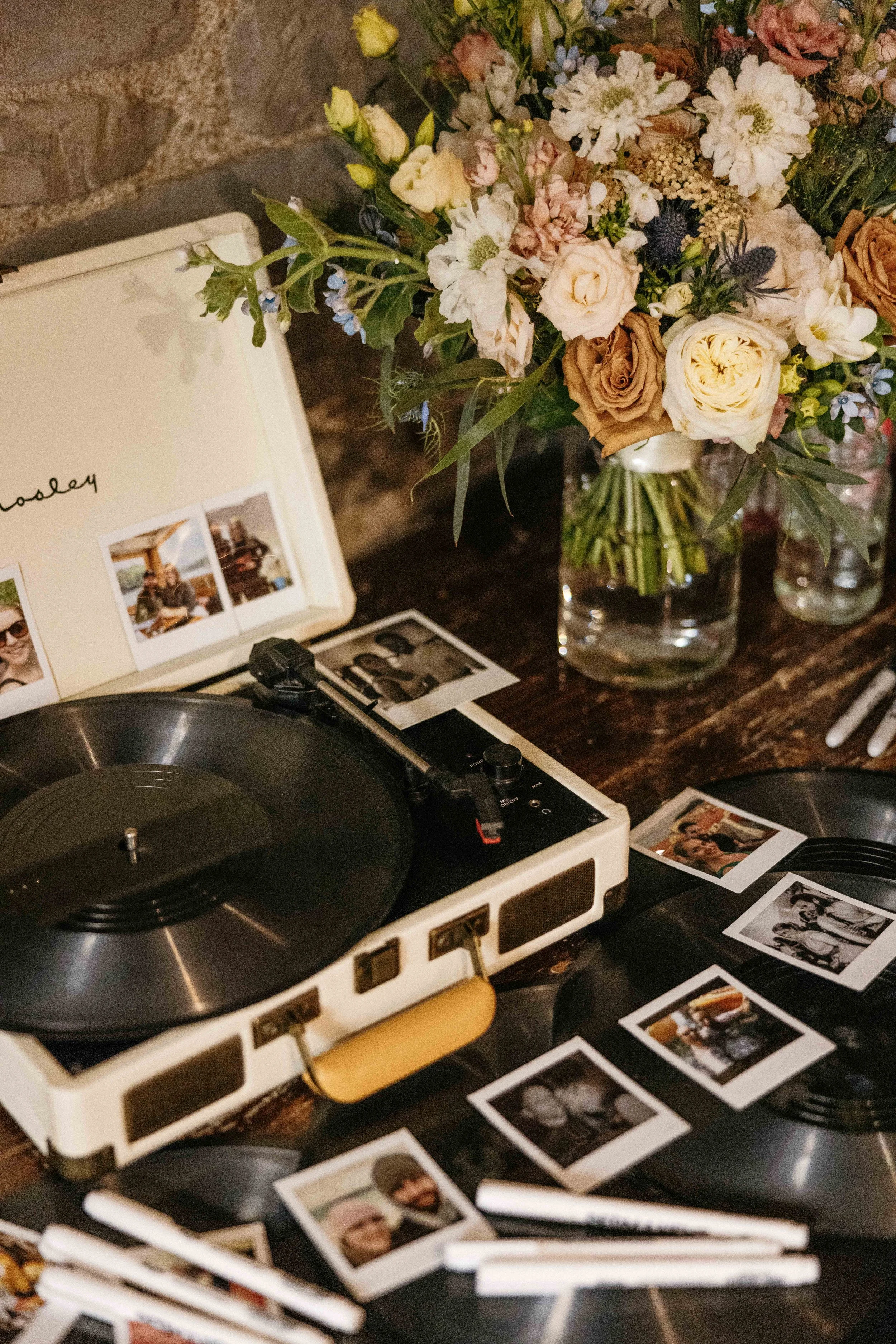 A vintage turntable on a wooden table surrounded by Polaroid photos and a large bouquet of assorted flowers in a glass vase.