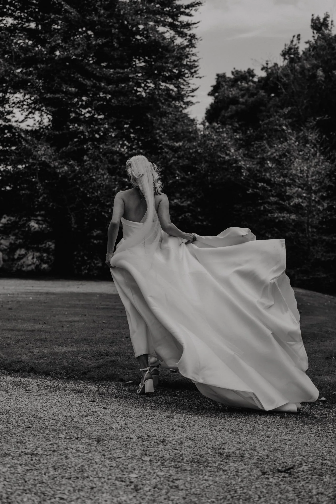 A woman in vintage attire is walking outdoors on a gravel path, wearing a long flowing dress, high heels, and has long, wavy hair. The scene is captured in black and white, with trees and cloudy sky in the background.