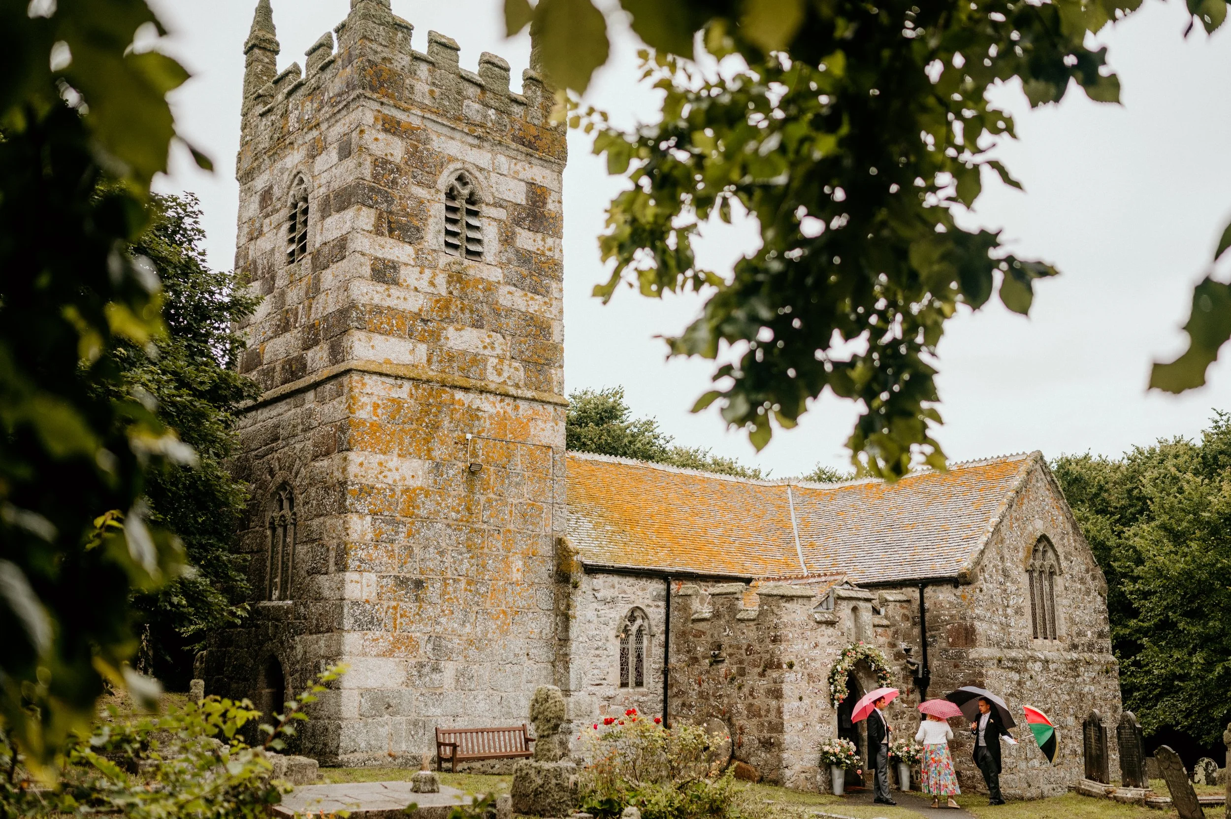 A group of people standing outside a historic stone church with a bell tower, holding umbrellas on a cloudy day.