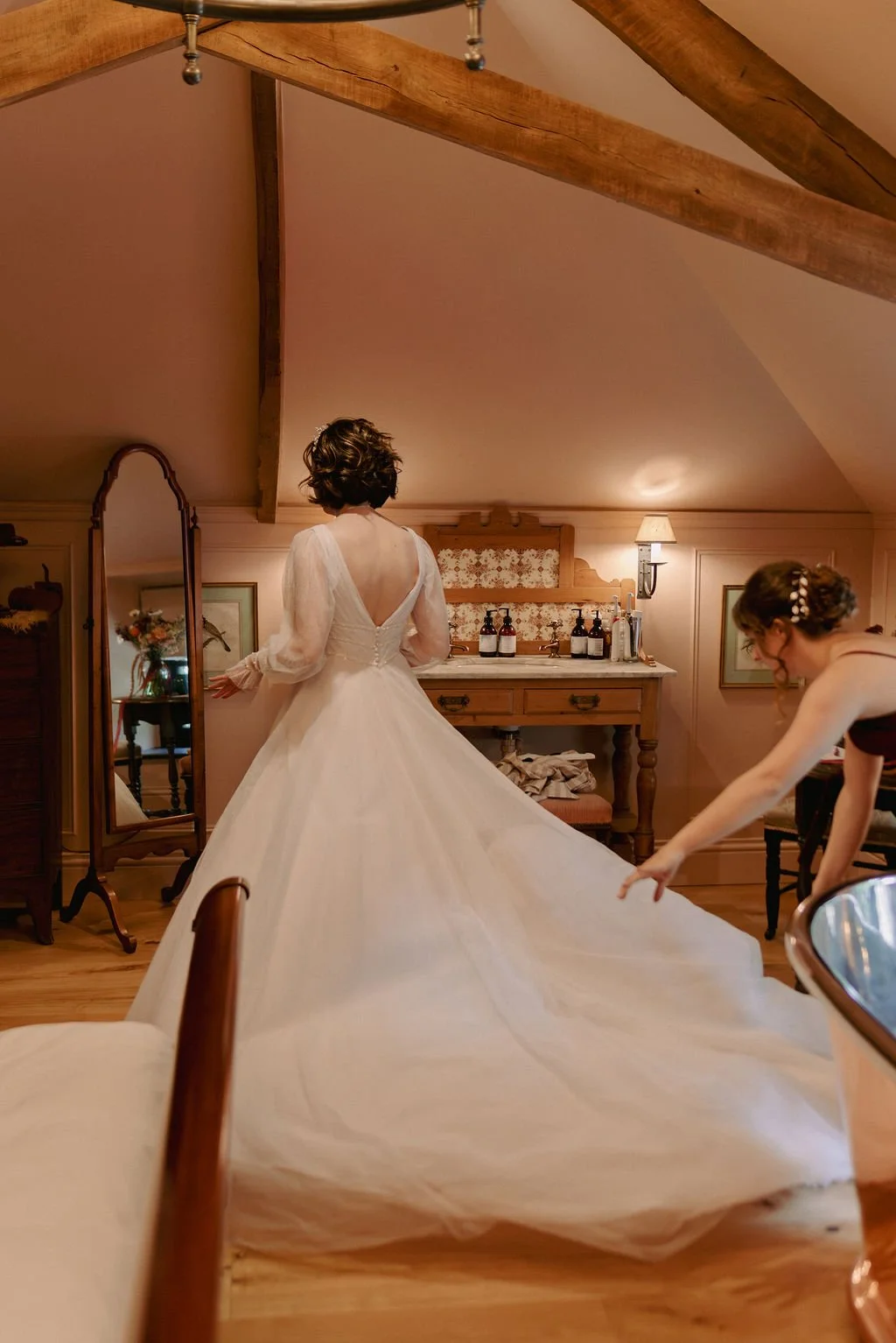 A bride in a wedding dress with a long train is getting ready in a cozy room while a woman helps arrange the train.