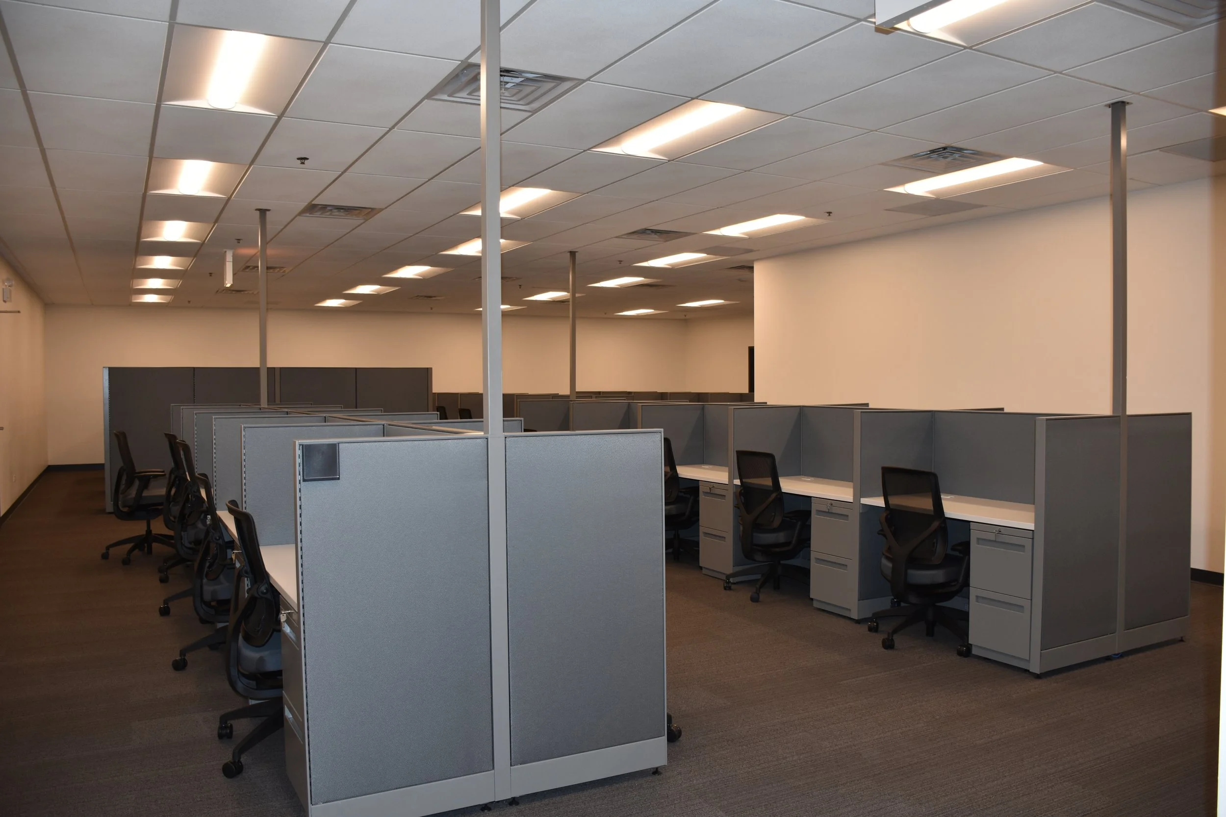 Empty office cubicles with chairs, fluorescent overhead lighting, and beige walls.