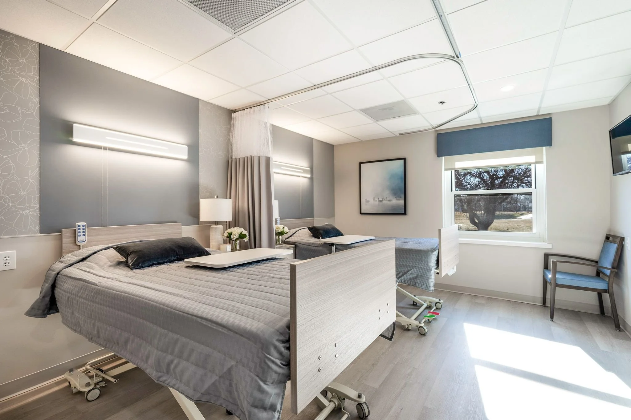 Hospital room with two hospital beds, bedside tables with lamps and flowers, a painting on the wall, window with tree outside, and medical equipment.