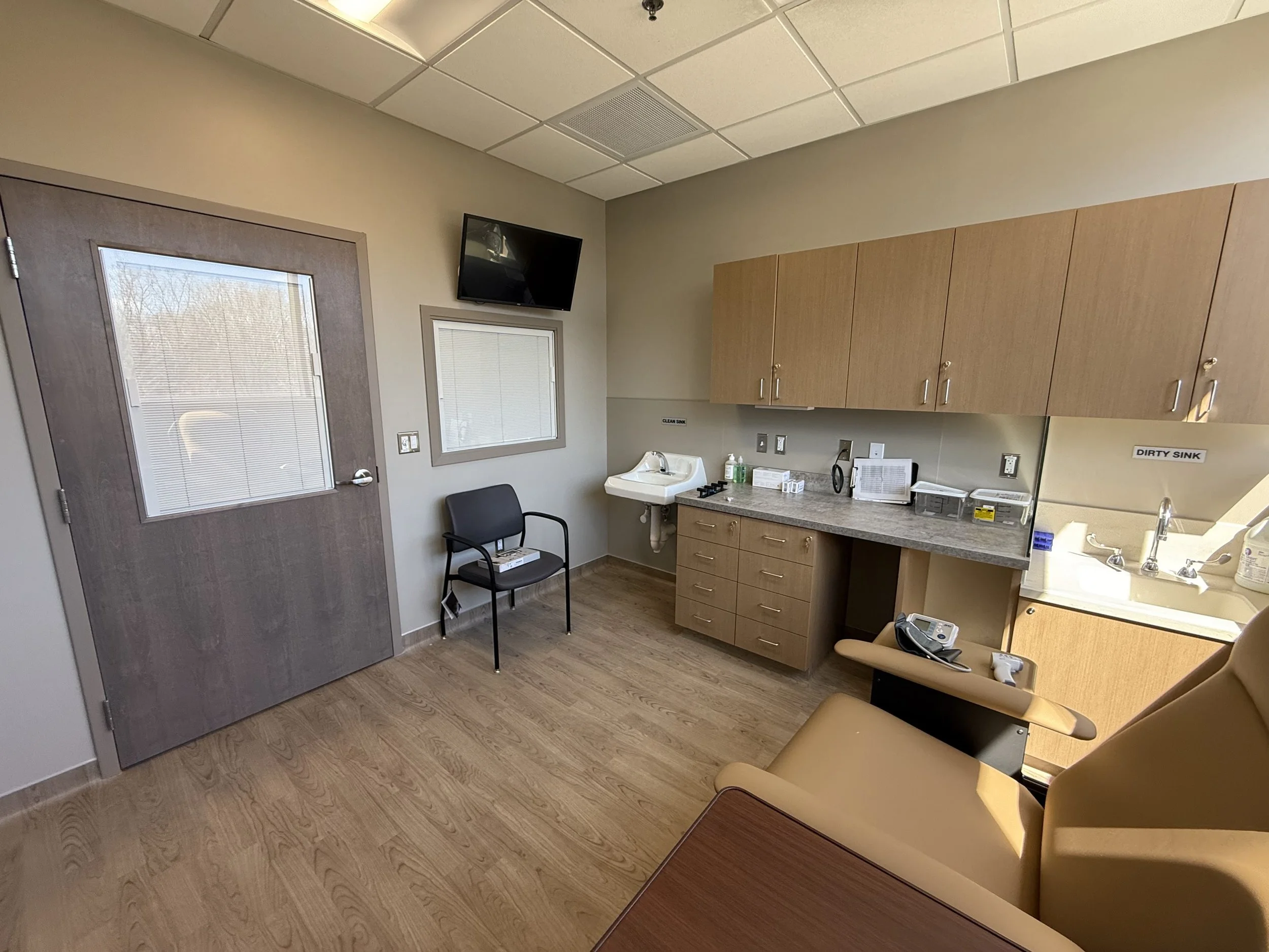 A medical examination room with a door, a chair, a countertop with medical equipment, cabinets, a sink, and a wall-mounted TV.