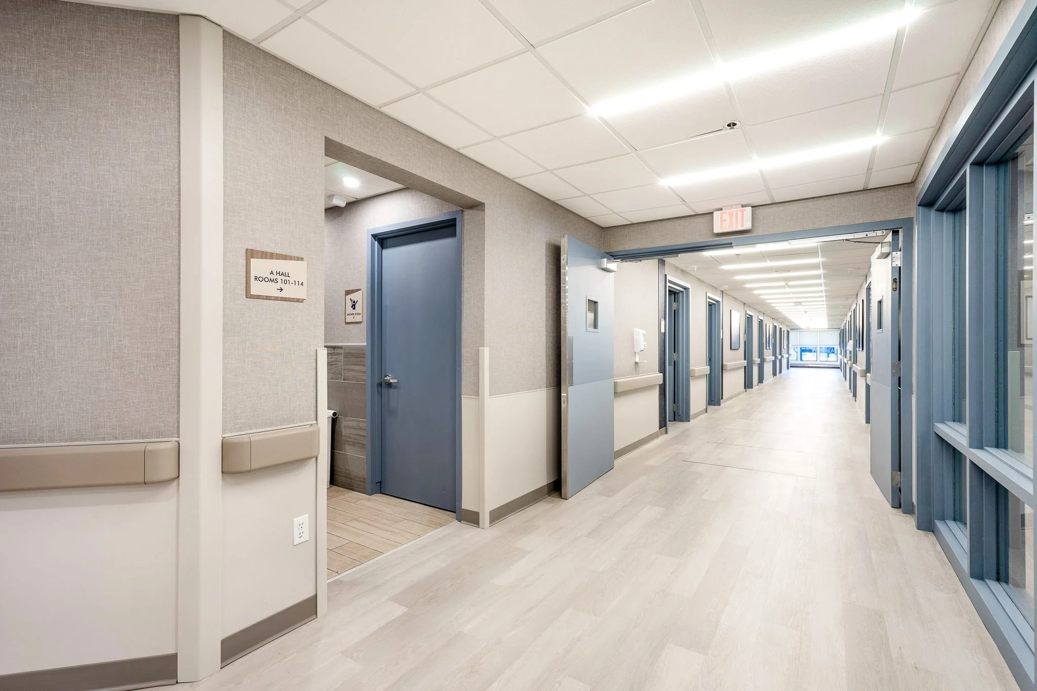 A hospital corridor with multiple examination rooms featuring blue doors, an open door revealing a bathroom, and signs indicating room numbers 101-114 and a water closet. The corridor is well-lit with ceiling lights and has a light-colored tiled floor.