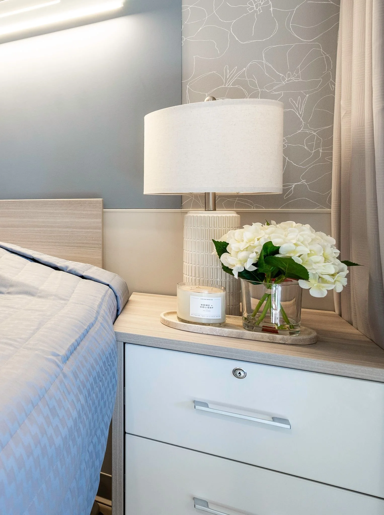 A bedroom nightstand with a white table lamp, a glass vase with white flowers, and a candle, next to a bed with a light-colored quilt and a wooden headboard. The wall behind has light-colored wallpaper with a floral pattern.