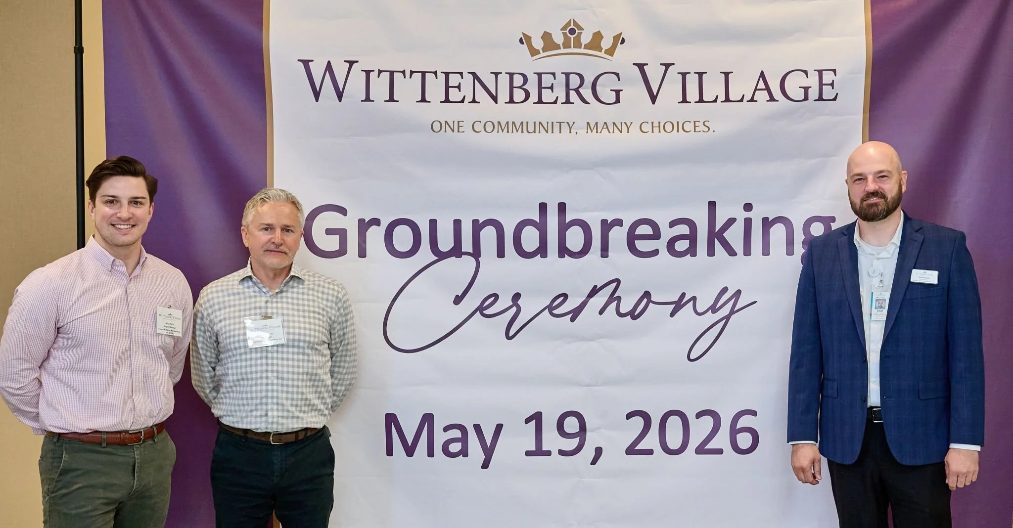 Three men standing in front of a large banner at a groundbreaking ceremony. The banner reads "Wittenberg Village, One Community, Many Choices, Groundbreaking Ceremony, May 19, 2026." The men are dressed in business casual attire and are smiling.