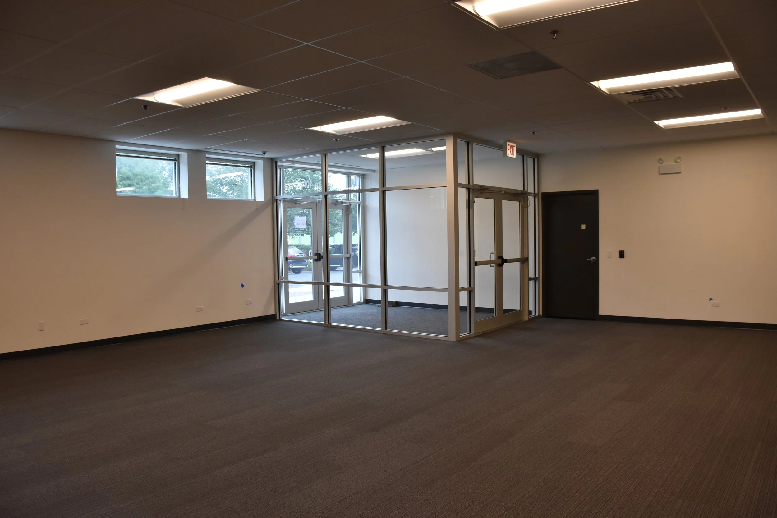 Empty room with dark carpet, beige walls, ceiling with fluorescent lights, exit sign, large glass windows, and a glass door.