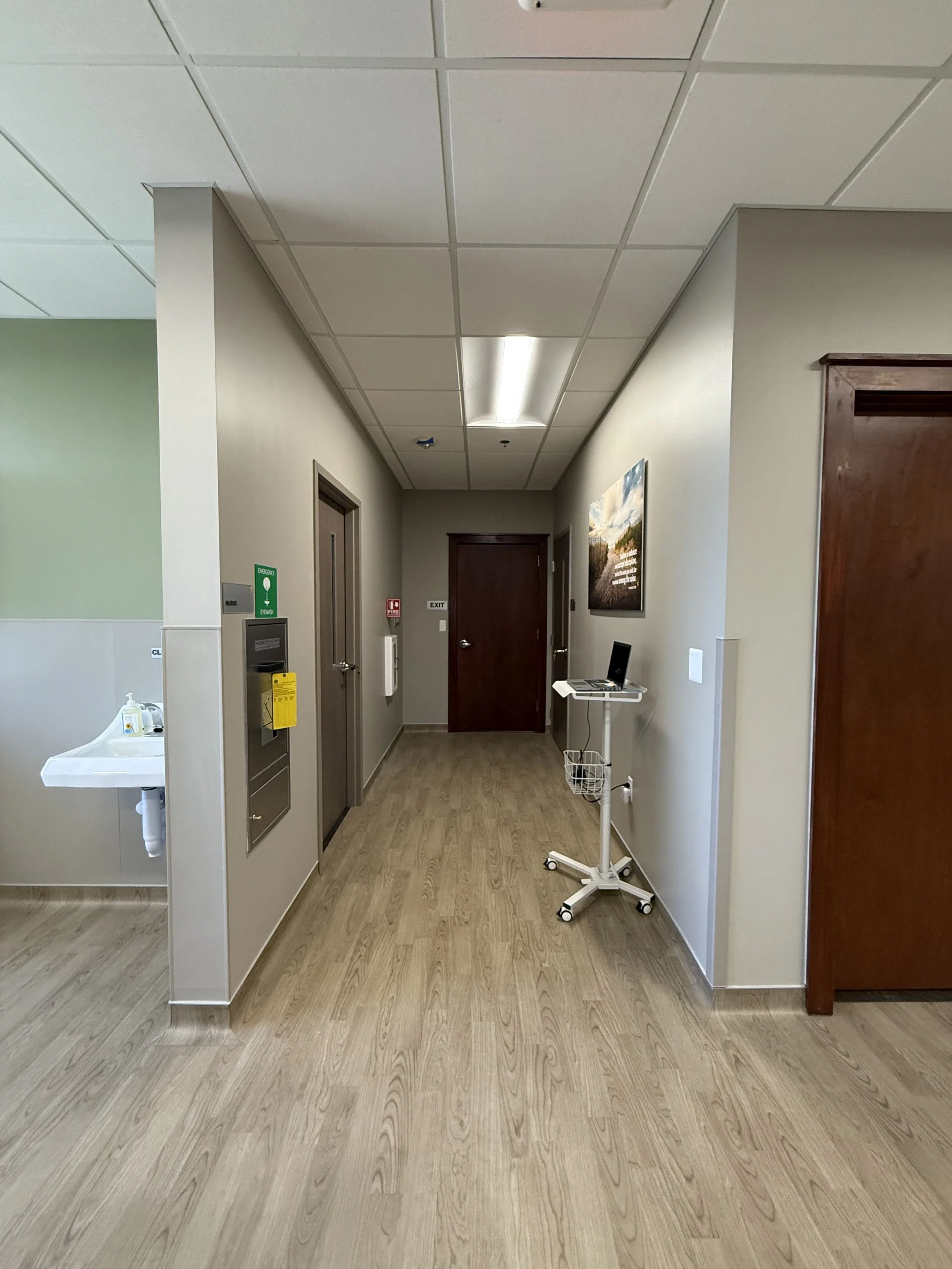 Hospital hallway with light wood flooring, beige walls, and a white ceiling with fluorescent lights. On the left side, there is a water sink with sanitizer and a green emergency exit sign. Toward the end of the hallway, there are closed doors, one with an 'Exit' sign, and a clipboard stand with a laptop and wheels on the right side. There is a framed landscape picture on the right wall.