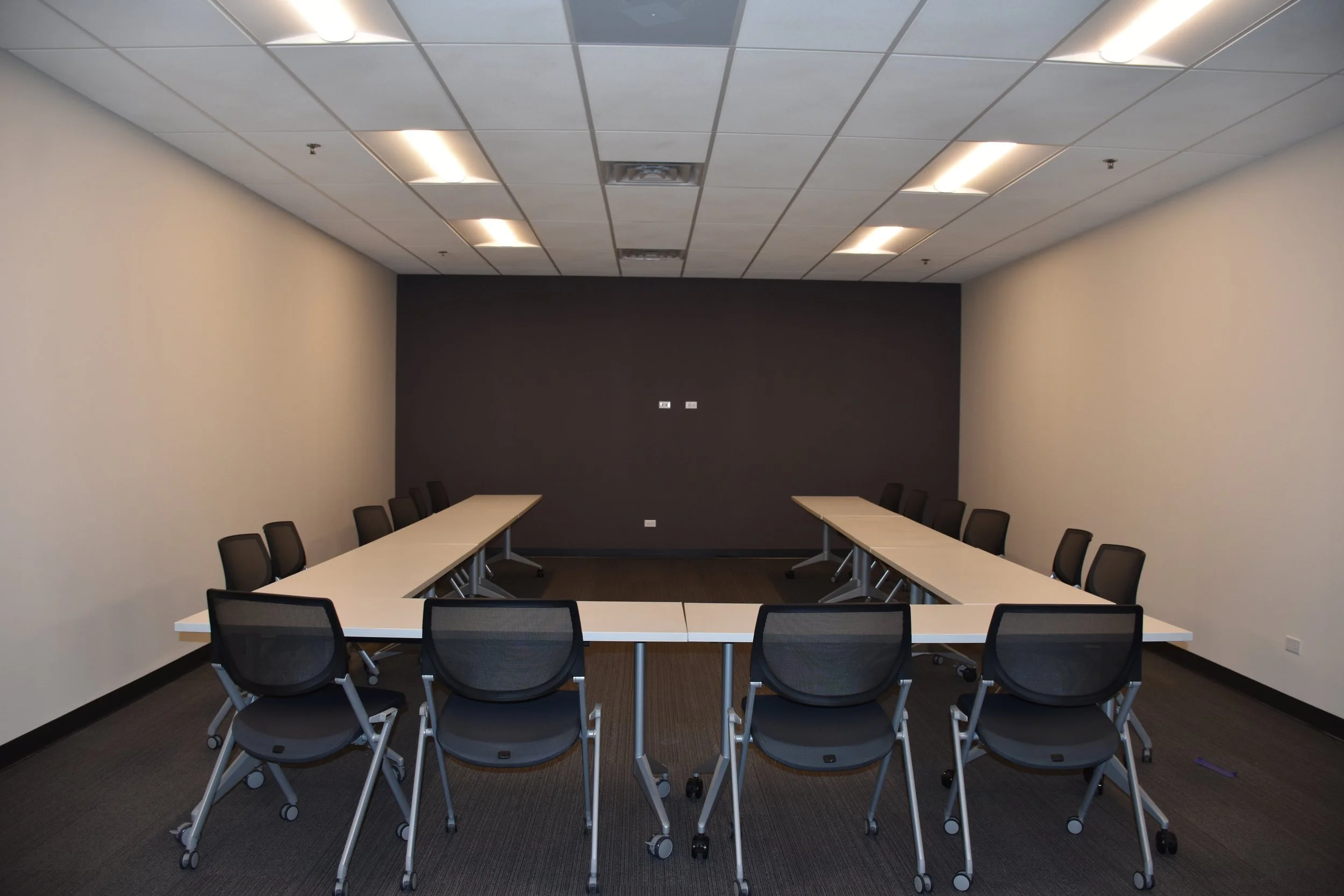 Empty conference room with a U-shaped arrangement of white tables and black chairs, beige walls, a dark accent wall, ceiling lights, and electrical outlets on the back wall.