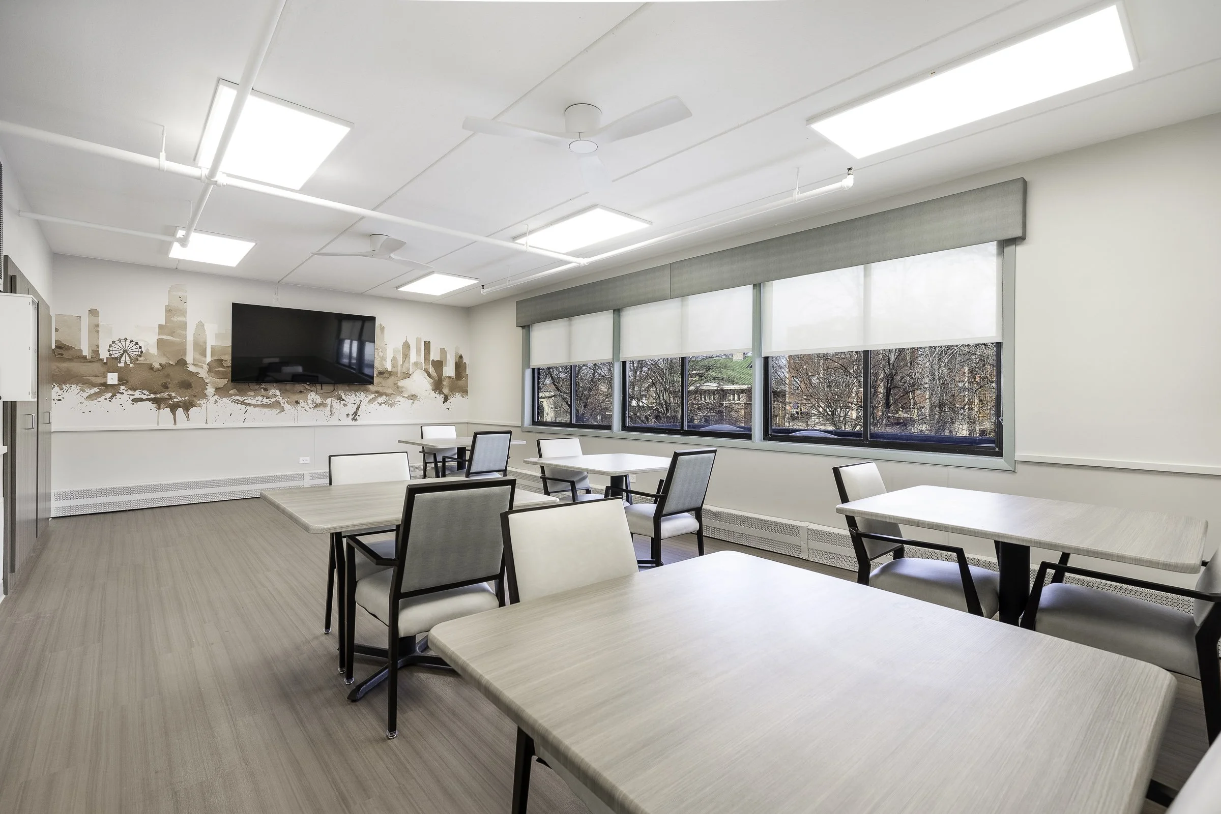 Empty classroom with white walls, large windows with blinds, and tables and chairs. A wall with a cityscape mural, a mounted flat-screen TV, and ceiling fans visible.