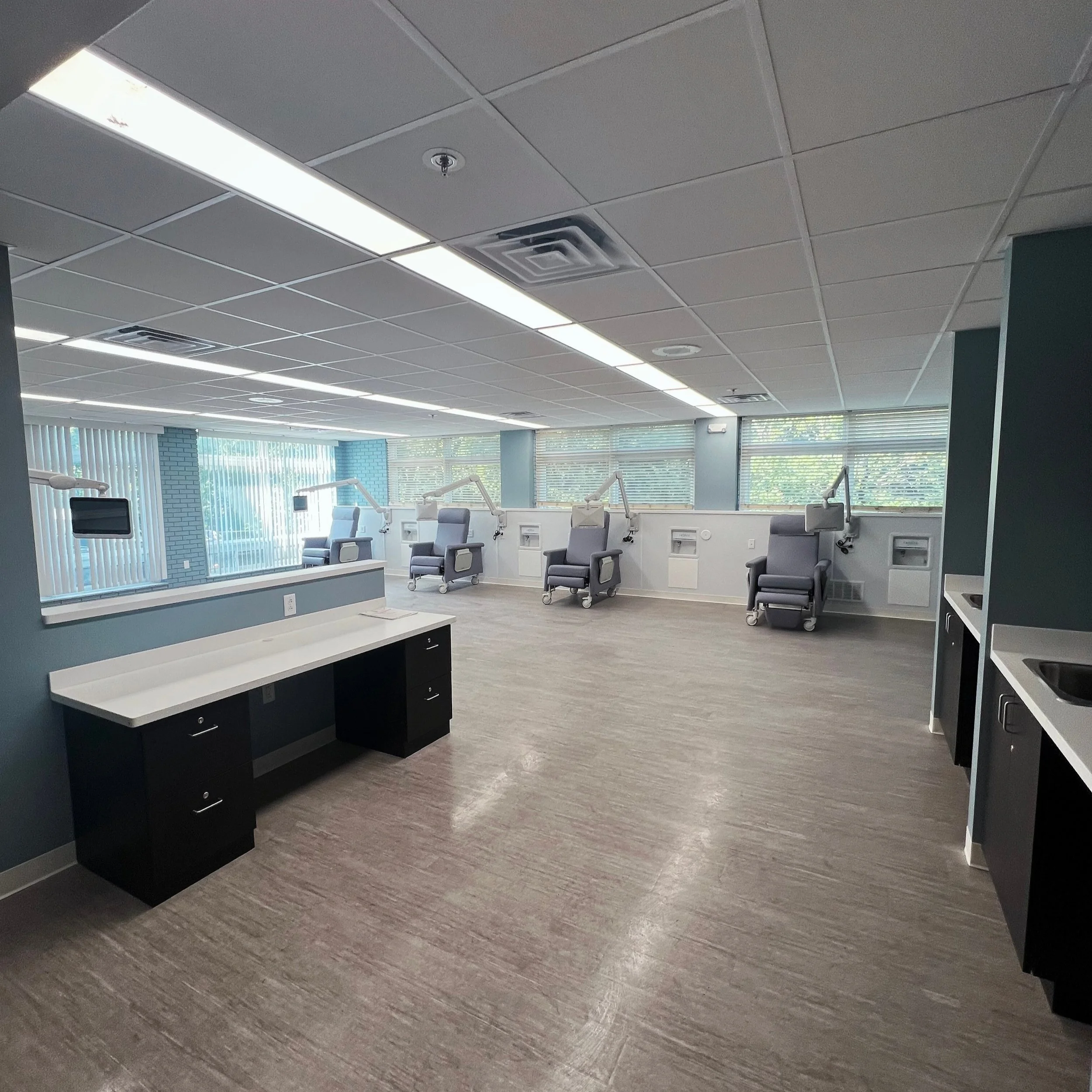 A line of four dialysis patient treatment stations, each with individual TVs on articulating arm-mounts and mocha-colored custom millwork cabinets and Corian countertops.