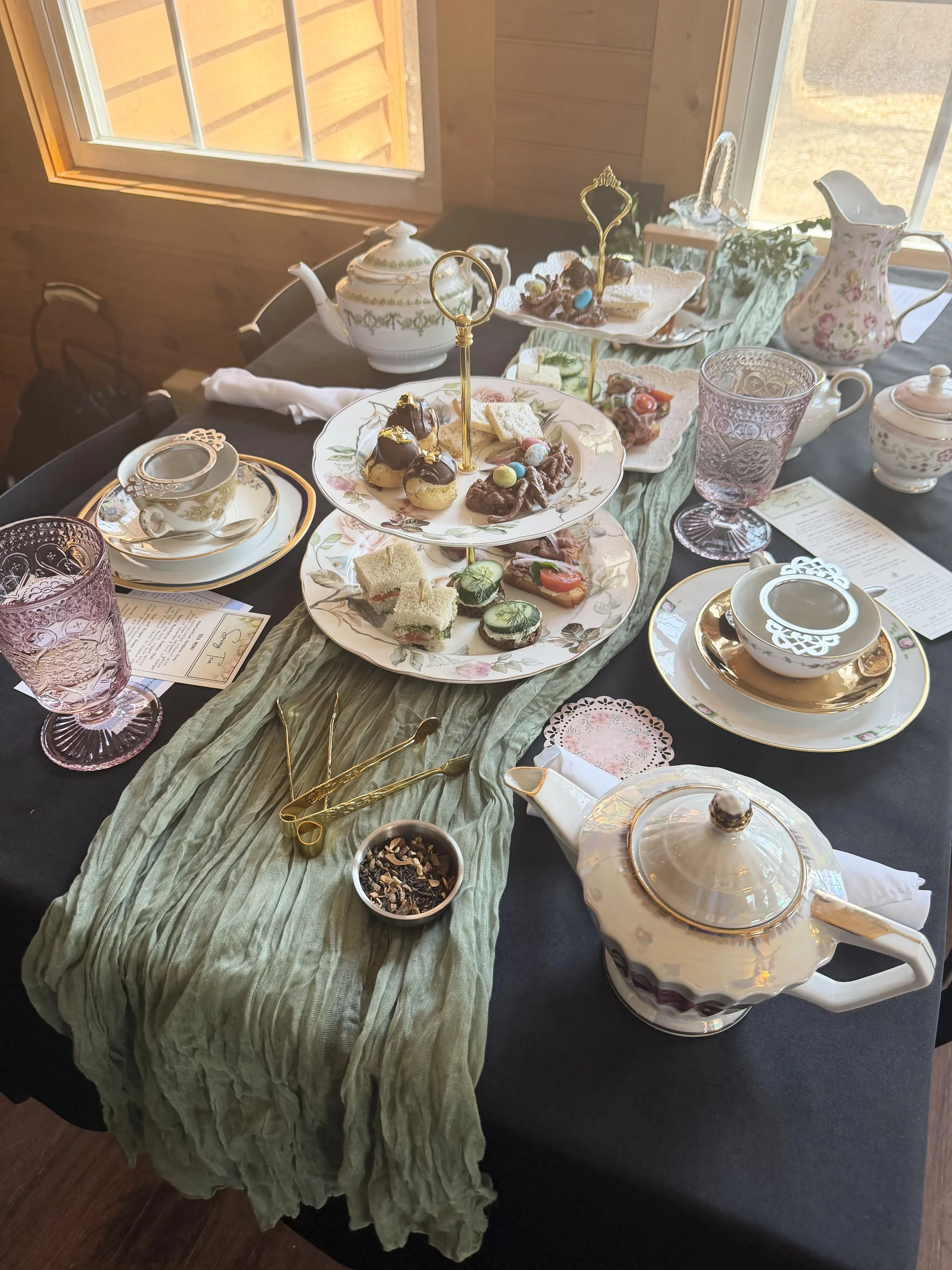 A table set for an elegant tea party with various plates of desserts, teacups, a teapot, water pitcher, and glasses, decorated with a green table runner and a menu placed on the table.