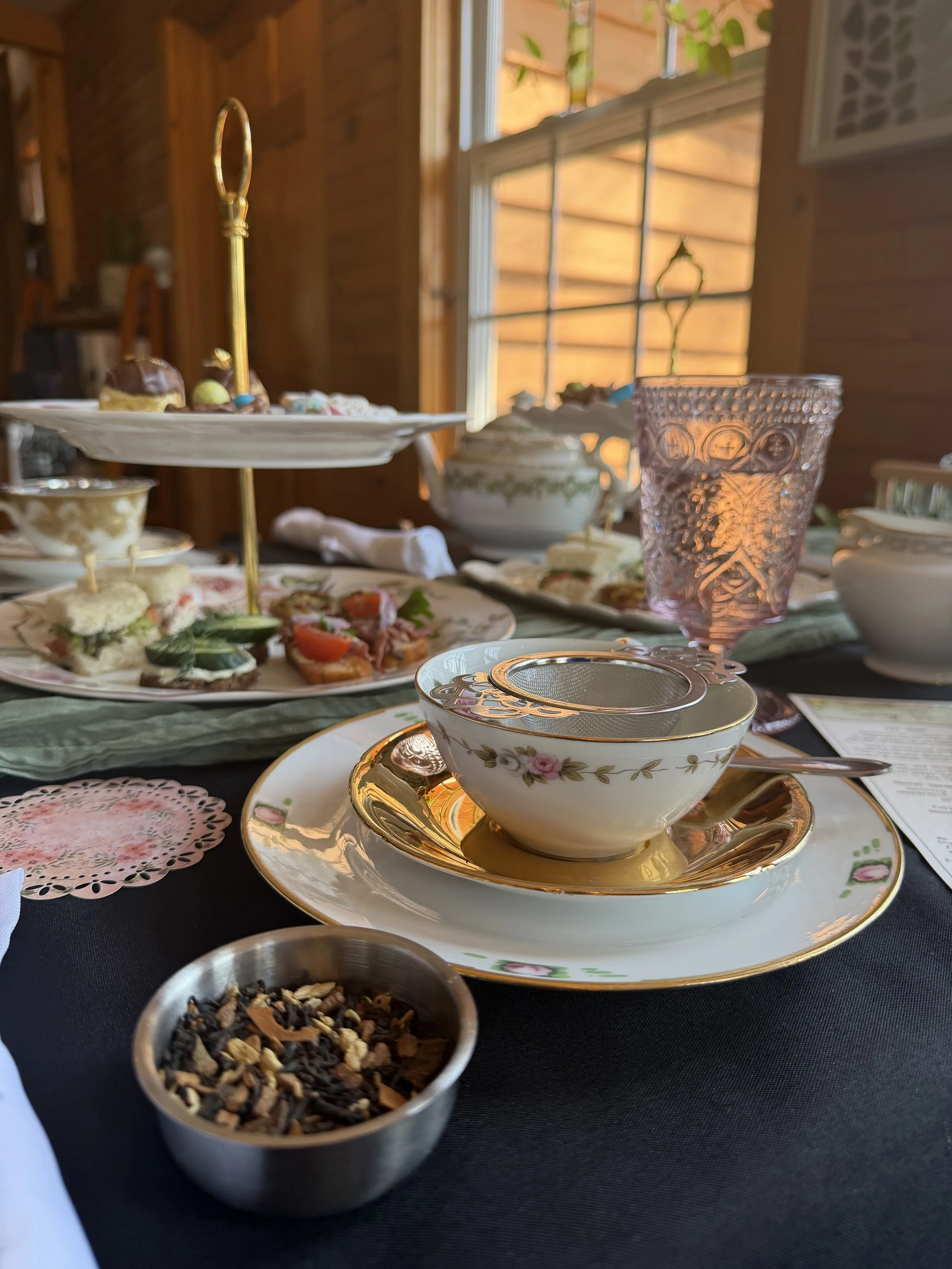 A table set for tea with fine china, a glass, a bowl of tea leaves, assorted finger foods, and a three-tier serving tray with desserts, in a room with wooden walls and a window letting in natural light.