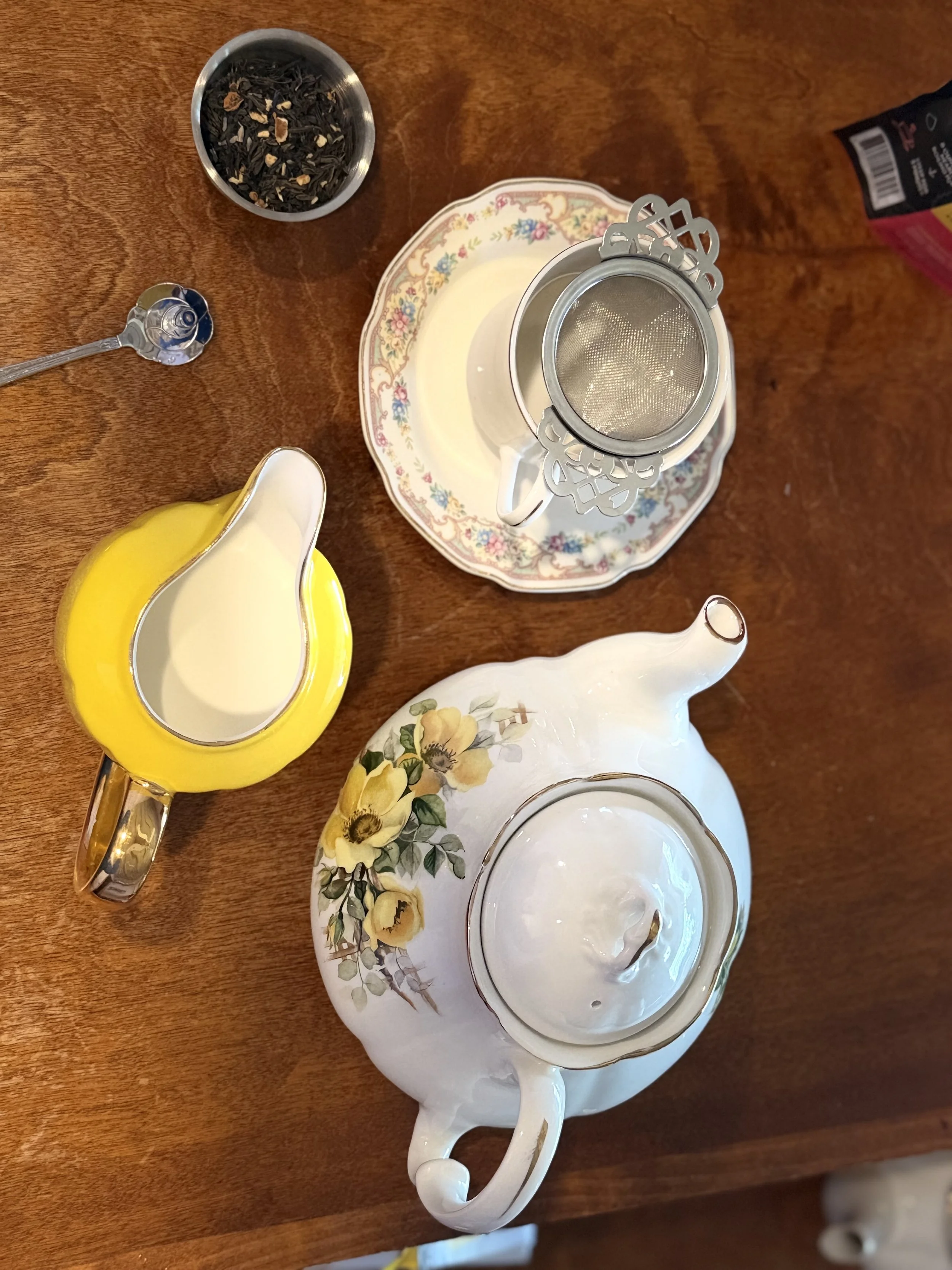 A collection of teaware and tea accessories on a wooden table, including a tea strainer, a yellow creamer, a floral teapot, and a floral teacup with milk.