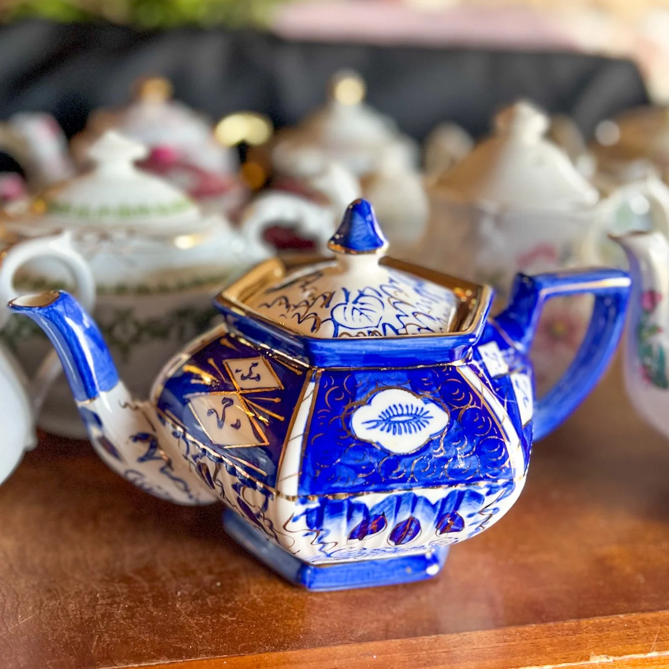 A decorative blue and white ceramic teapot with intricate gold detailing, placed on a wooden surface with blurred porcelain teapots in the background.