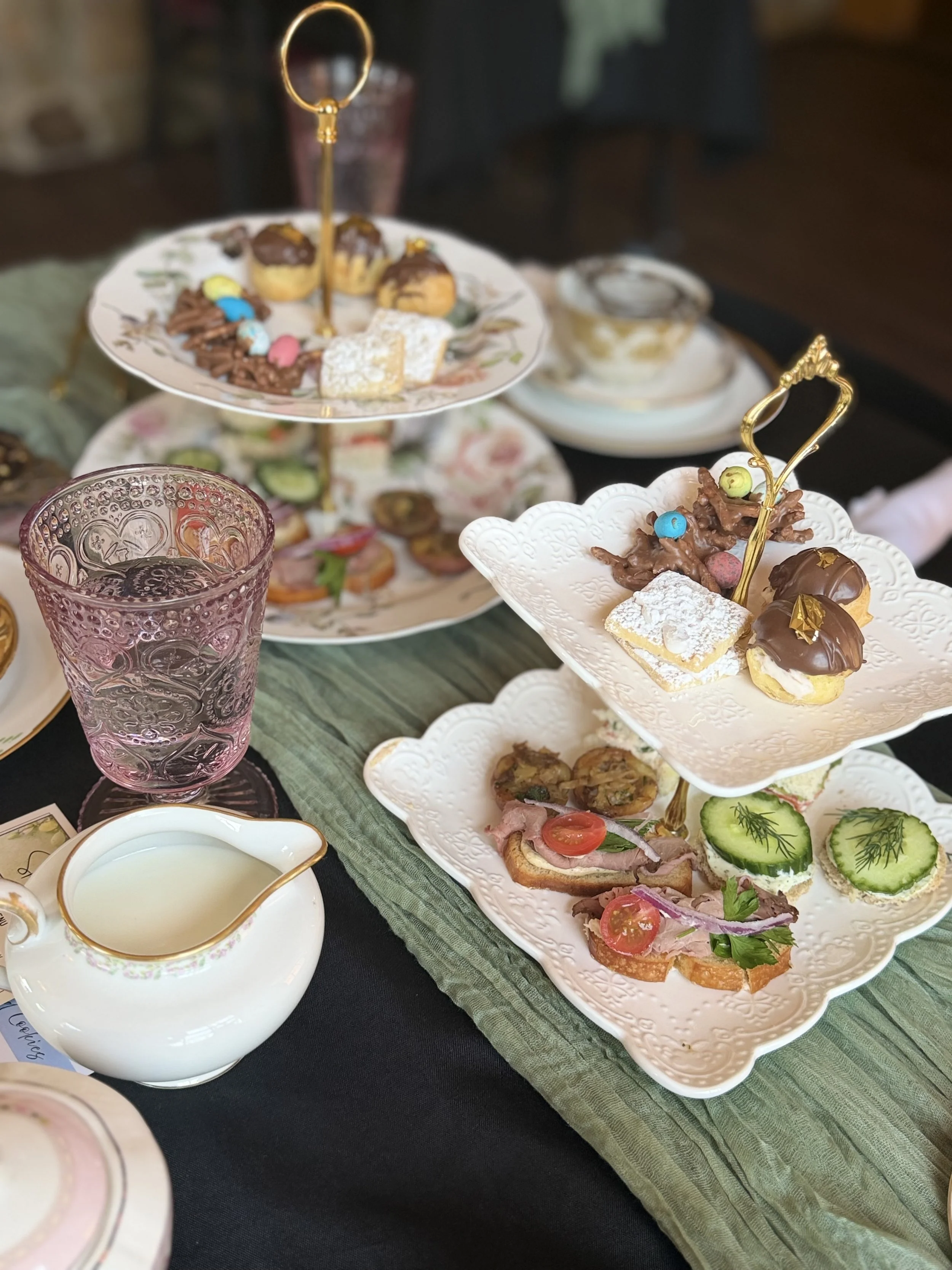 A multi-tiered tray filled with assorted desserts, including chocolate-covered treats, cookies, and small pastries, on a table with tea, a pink glass, and decorative plates.