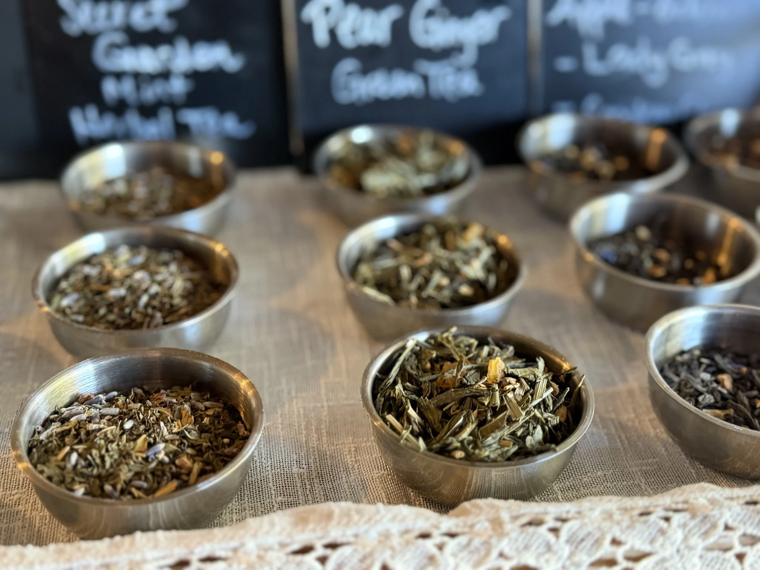 Metal bowls filled with various dried herbs and teas on a table with a chalkboard menu in the background.