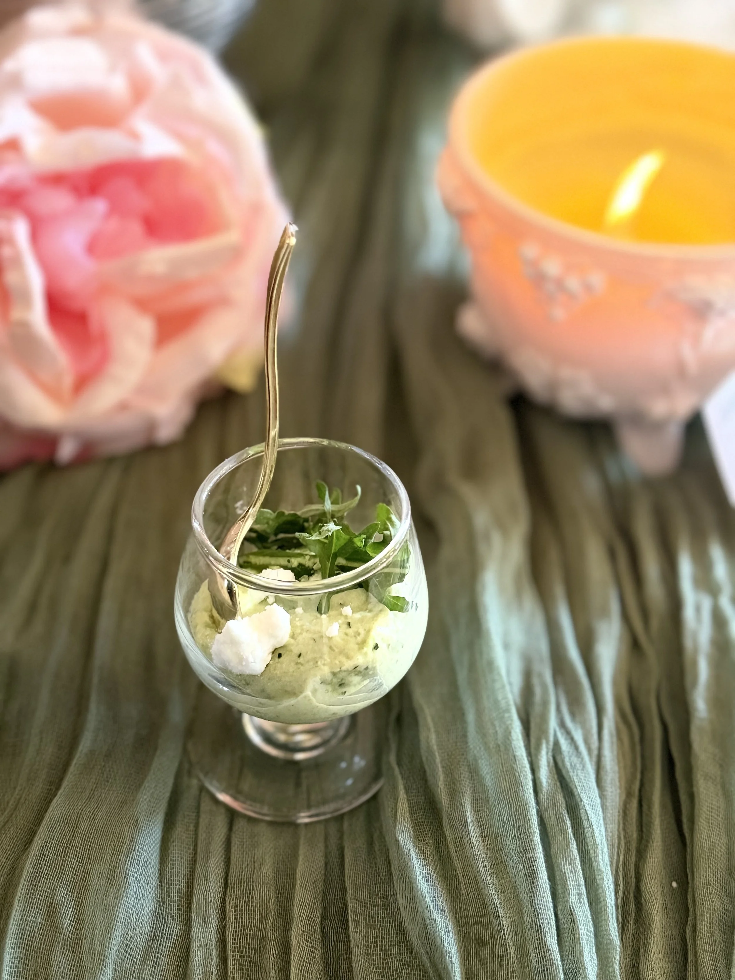 A small glass jar filled with green herbs, topped with white cheese or cream, on a textured green tablecloth, with blurred pink candles in the background.