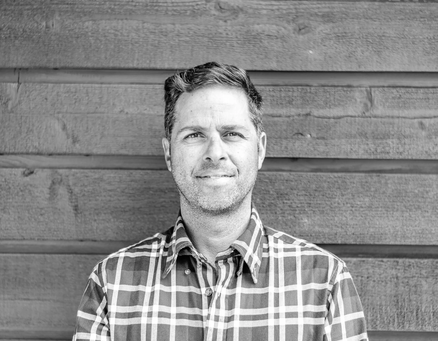 Black and white portrait of a man with short dark hair, wearing a plaid shirt, standing in front of a wooden wall, smiling slightly.