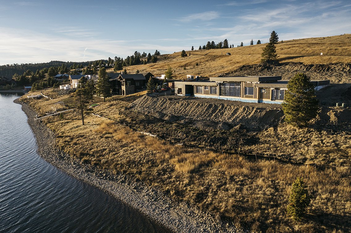 Residential houses under construction on a hillside near a lake with a rocky shoreline and sparse trees, under a partly cloudy sky.