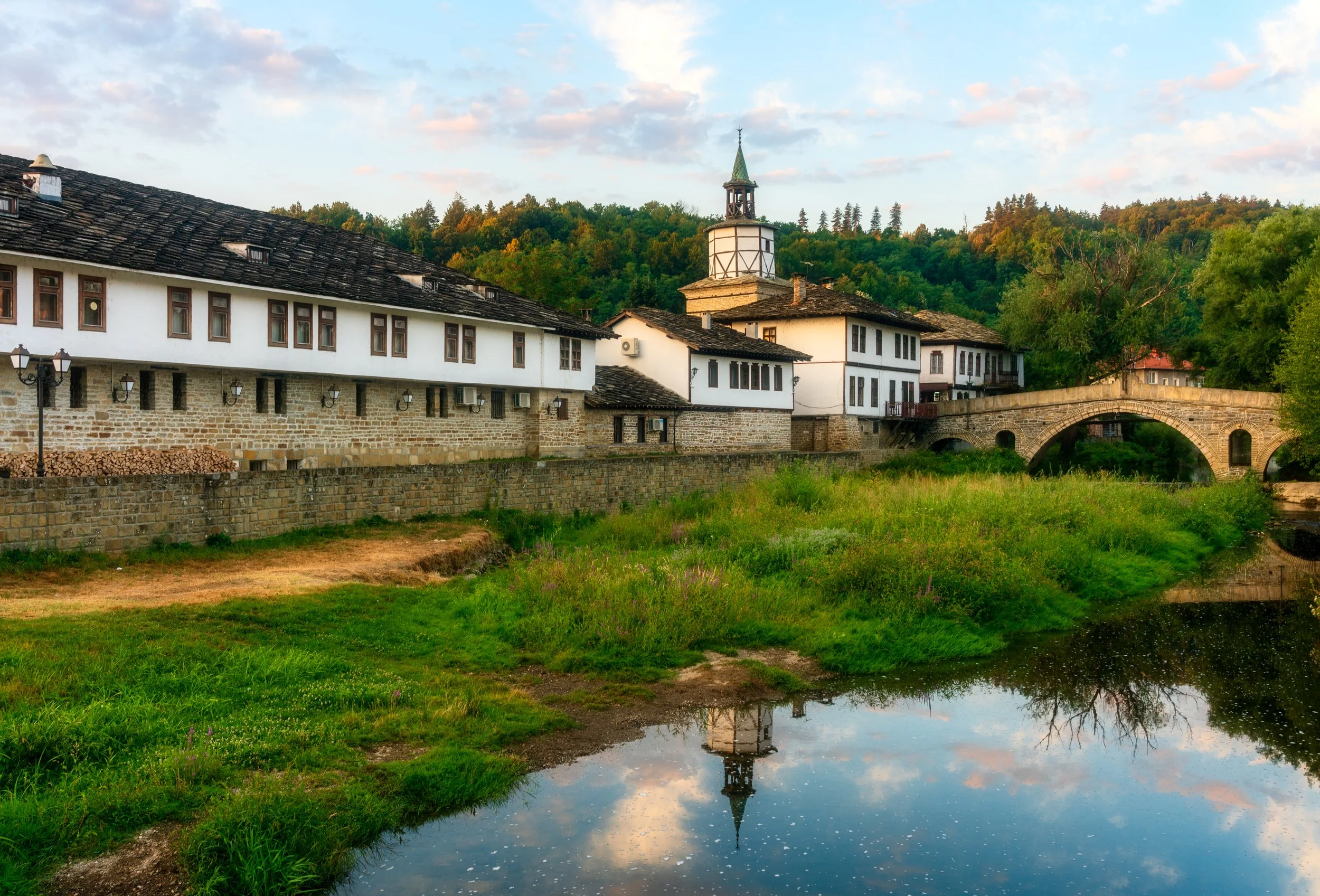 Evening view of the Revival-era architecture in Tryavna, Bulgaria, featuring traditional whitewashed houses with stone roofs and the iconic Clock Tower of Tryavna reflected in the calm river. The arched stone bridge and lush greenery add to the seren
