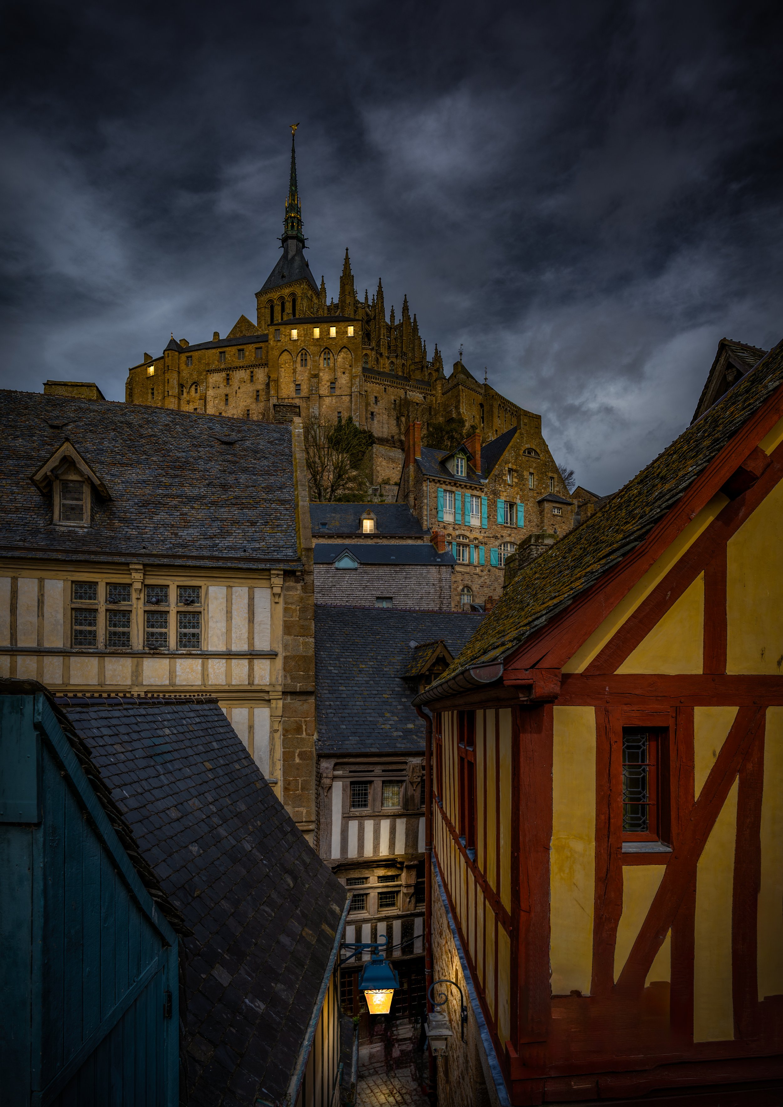 A dramatic view of Mont Saint-Michel Abbey rising above the medieval streets at dusk in Normandy, France. Fine art travel and architectural photography capturing one of France's most iconic UNESCO World Heritage Sites under stormy evening skies.