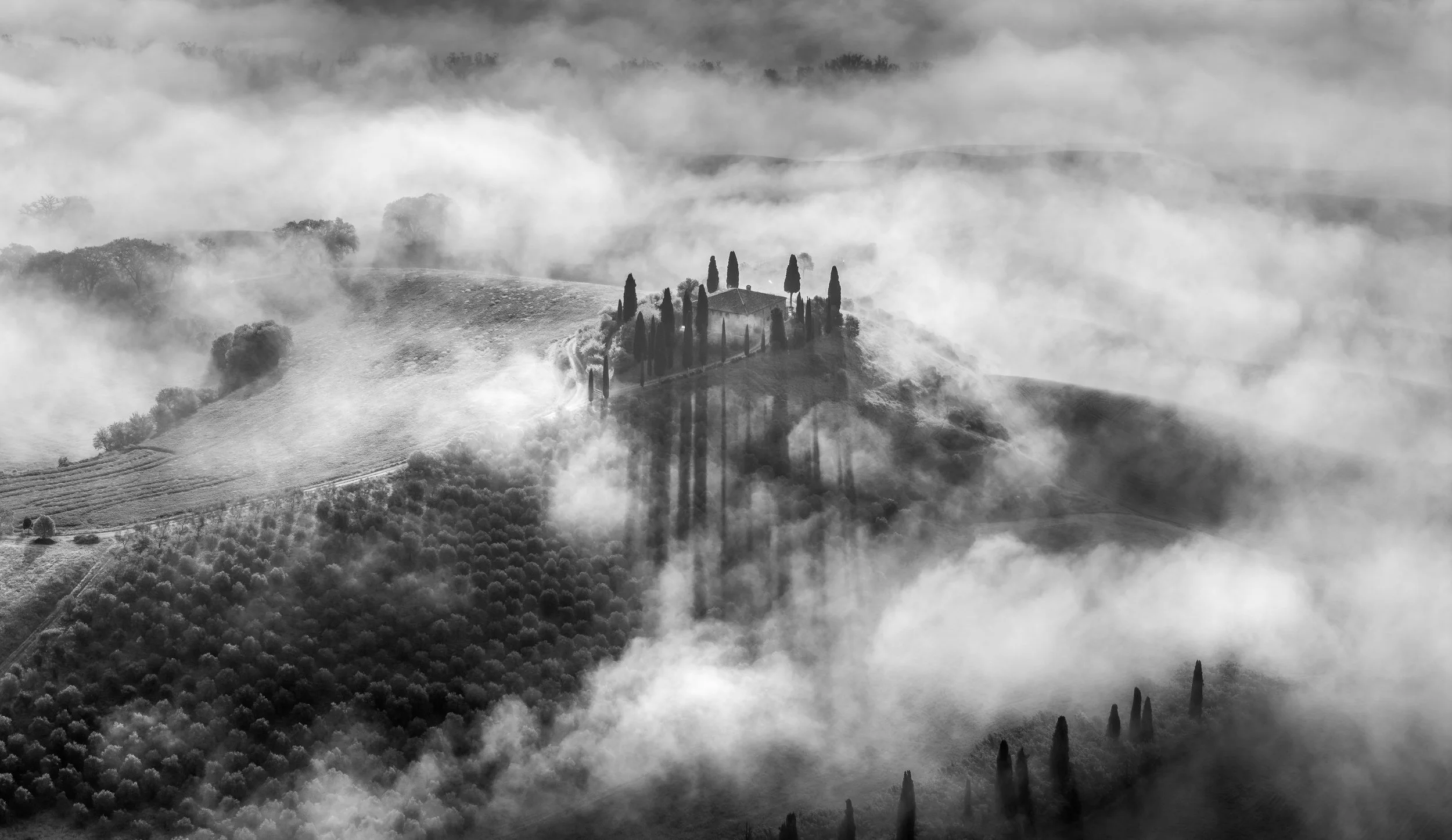 Atmospheric black and white aerial photograph of Tuscany’s rolling hills, where a secluded farmhouse and tall cypress trees rise through dense morning fog. The monochrome tones enhance the dramatic light and shadow, capturing the timeless beauty of t