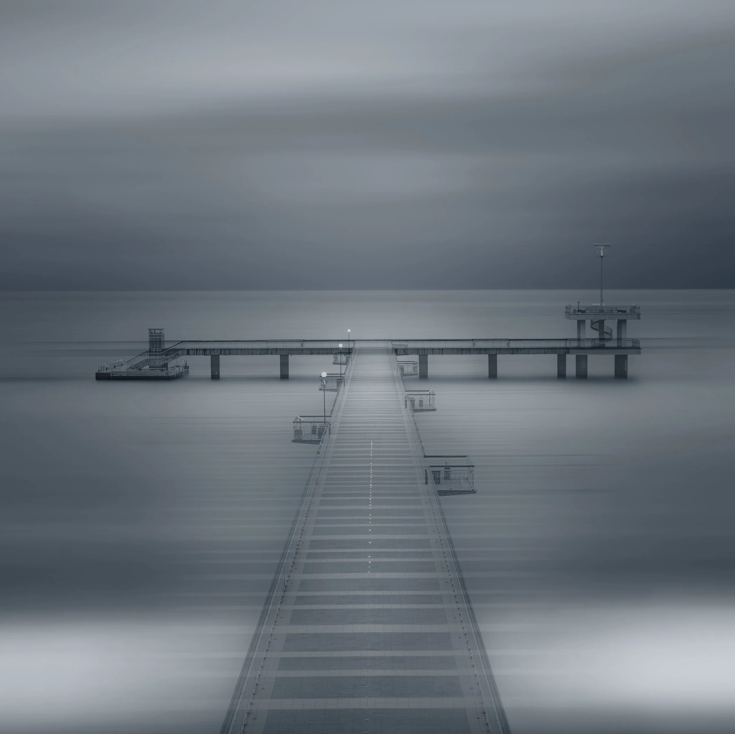 Minimalist long exposure photograph of a pier extending into the calm waters of the Black Sea under a moody, overcast sky. The smooth surface of the sea and soft tonal gradients create a serene, atmospheric composition focused on symmetry, depth, and