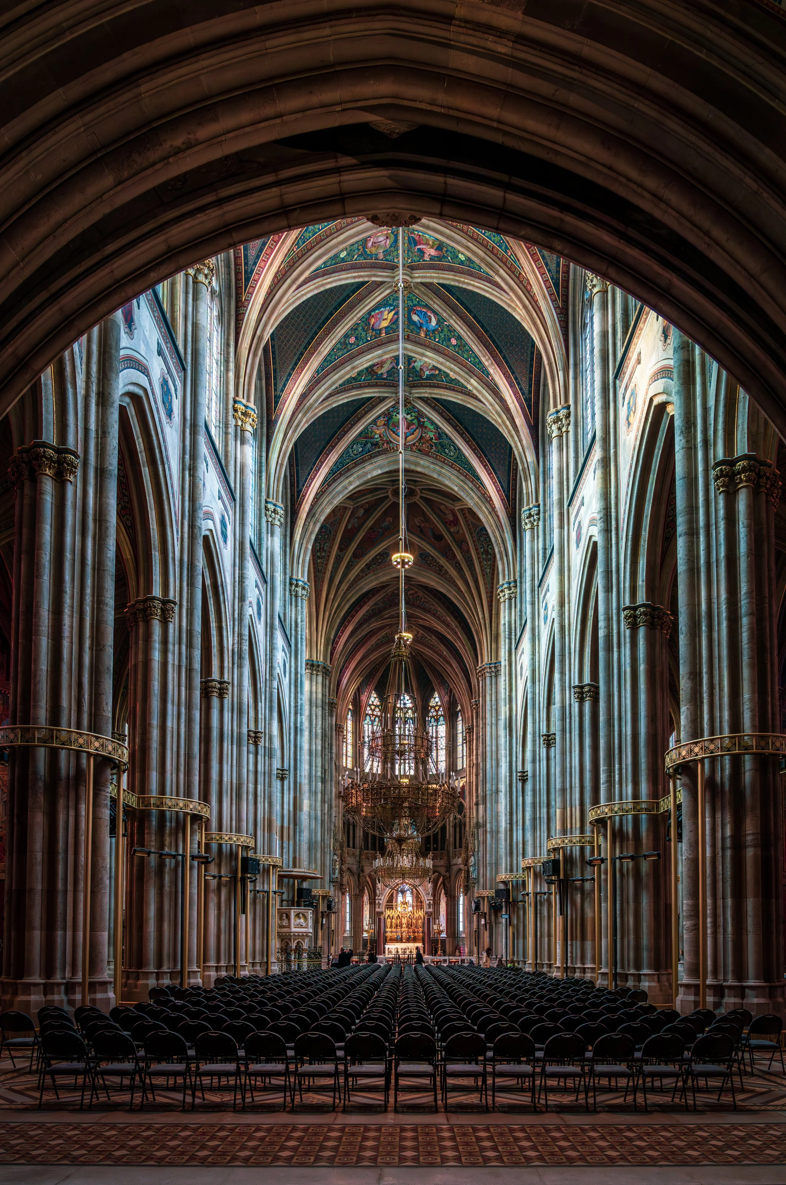 A stunning travel photography image of a grand Gothic cathedral interior in Vienna, showcasing towering vaulted ceilings, richly decorated arches, and intricate architectural details. Rows of chairs lead toward an illuminated altar, creating a powerf