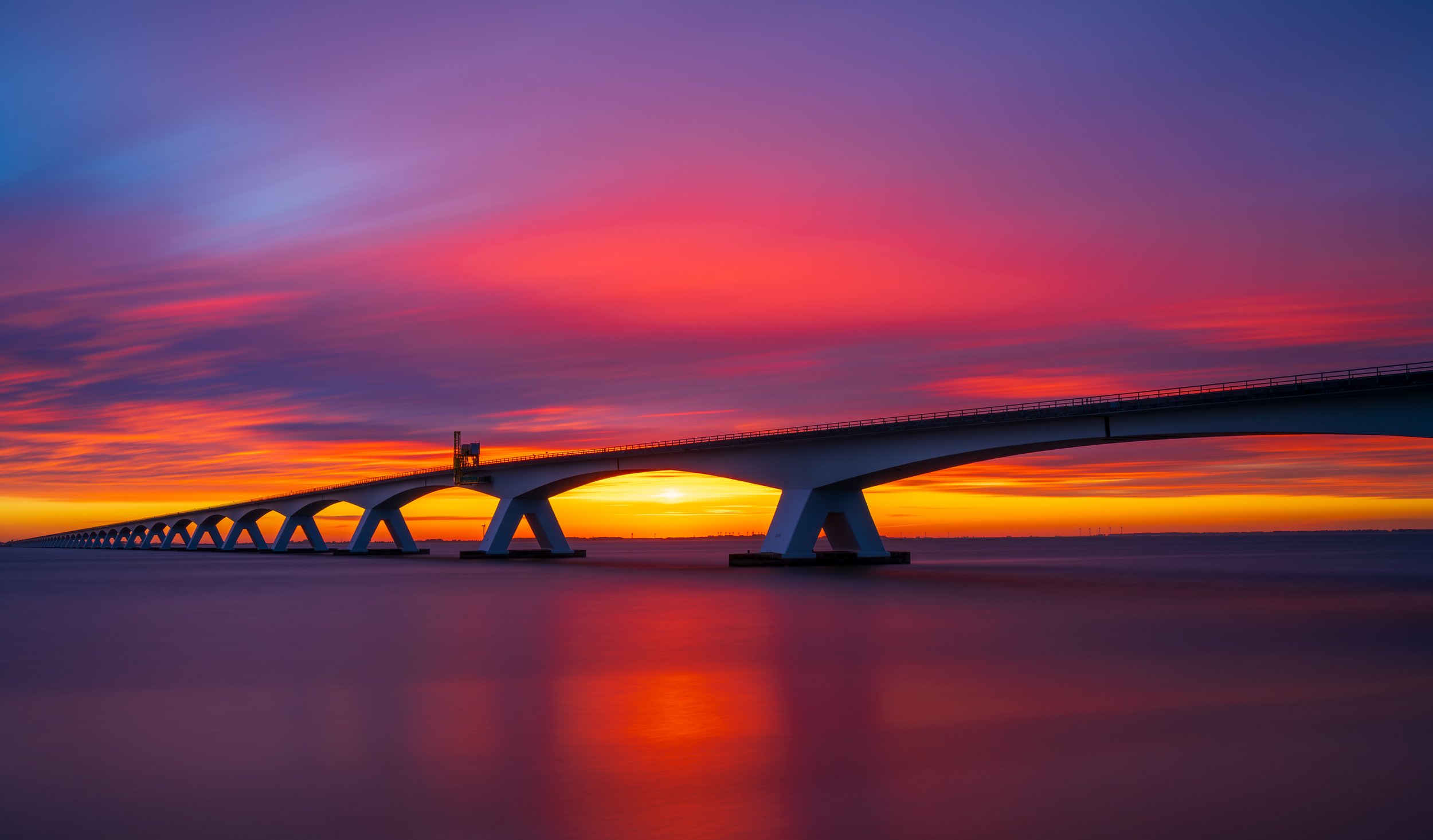 Long exposure sunset photograph of the Zeeland Bridge stretching across calm waters under a vibrant red and purple sky. The smooth reflections and bold colors create a dramatic fine art seascape, emphasizing symmetry, architecture, and the power of n