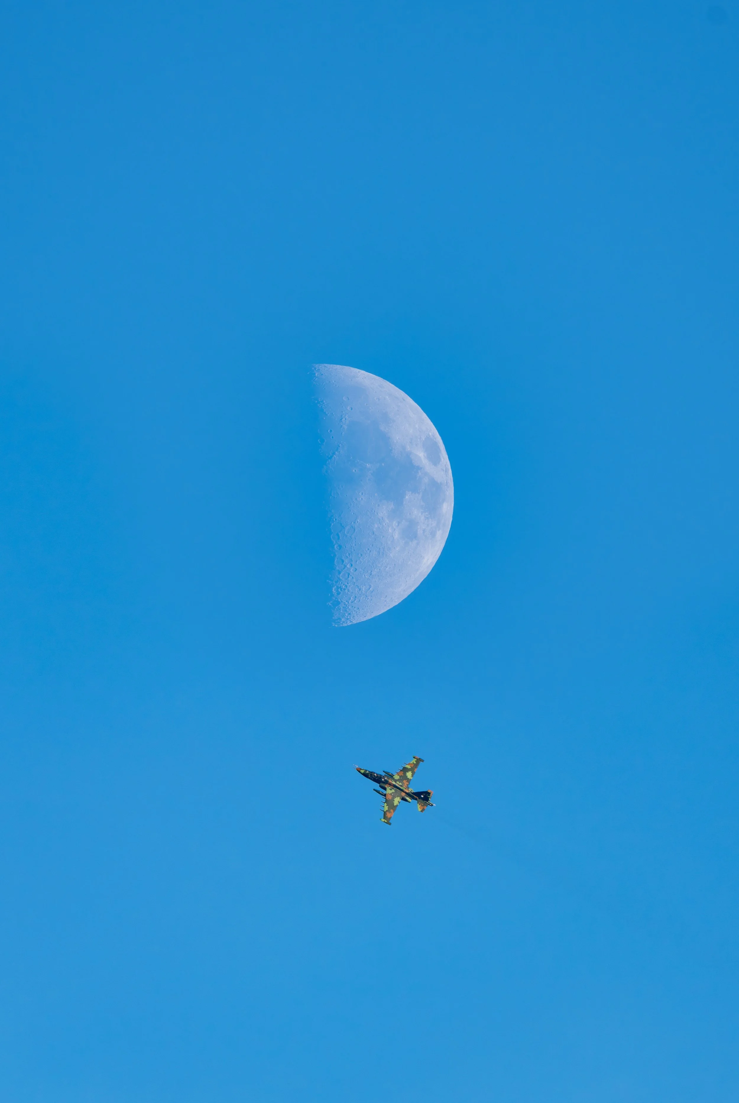 A fleeting moment captured against a vast, cloudless sky: a military aircraft cuts across the frame beneath a sharply defined daytime moon. The composition plays on scale and contrast-human motion set against a timeless celestial presence-creating a 