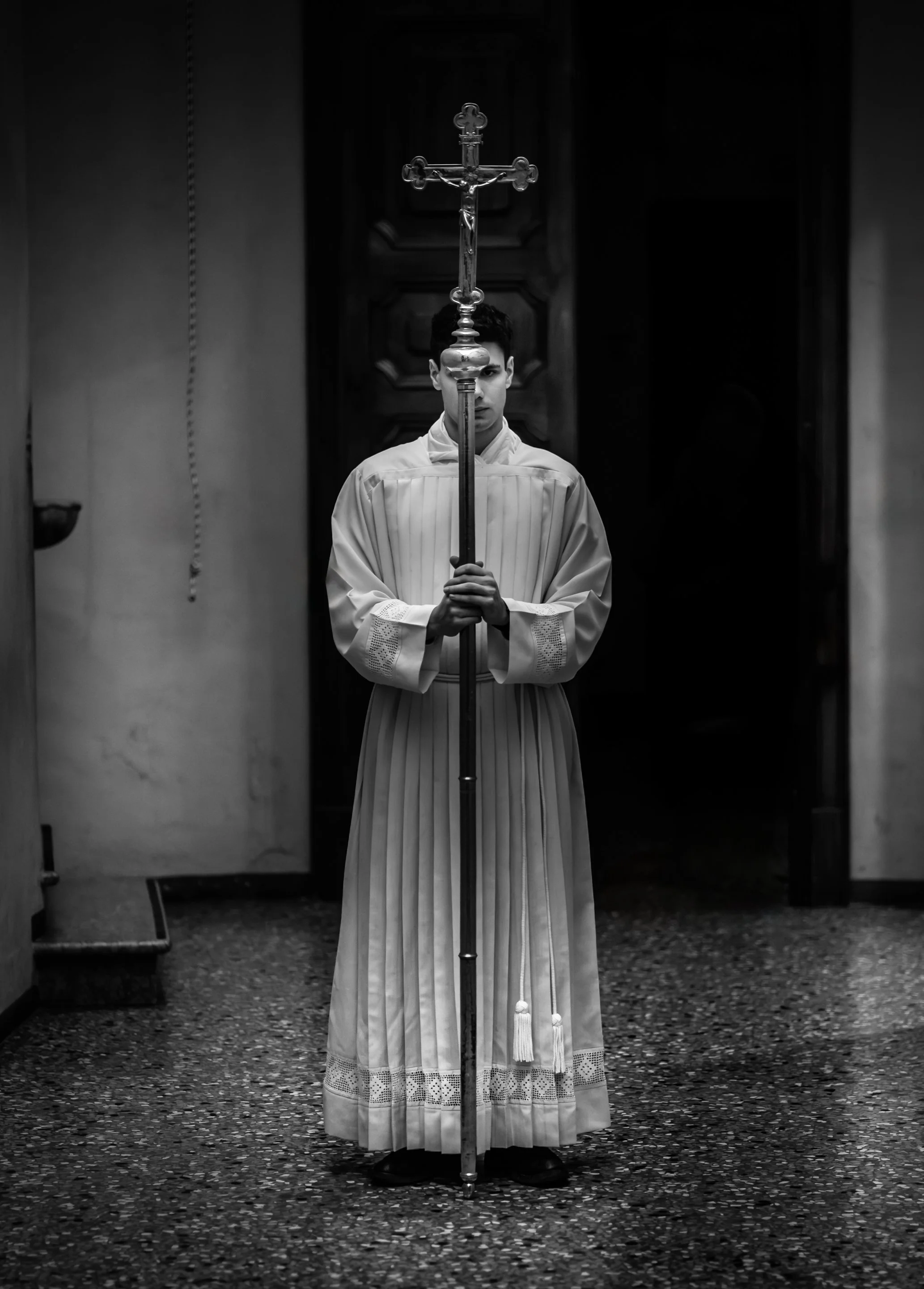 Black and white photograph of an altar server holding a processional cross inside a Catholic church.