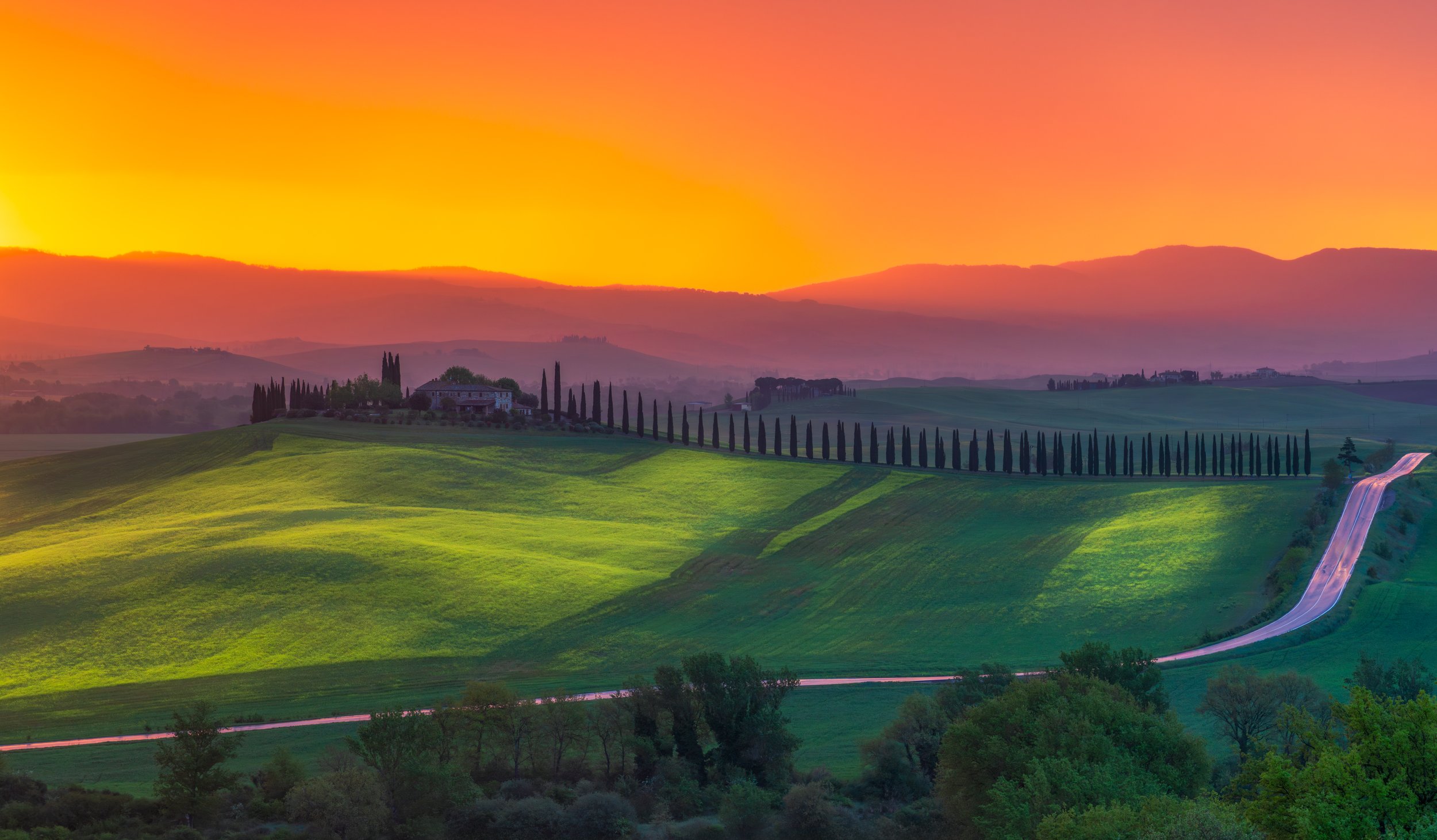 Stunning sunset landscape in Val d’Orcia, Tuscany, featuring rolling green hills, a traditional farmhouse, and a long cypress-lined road under a glowing orange sky. One of the most iconic views of the Italian countryside, perfect for travel inspirati