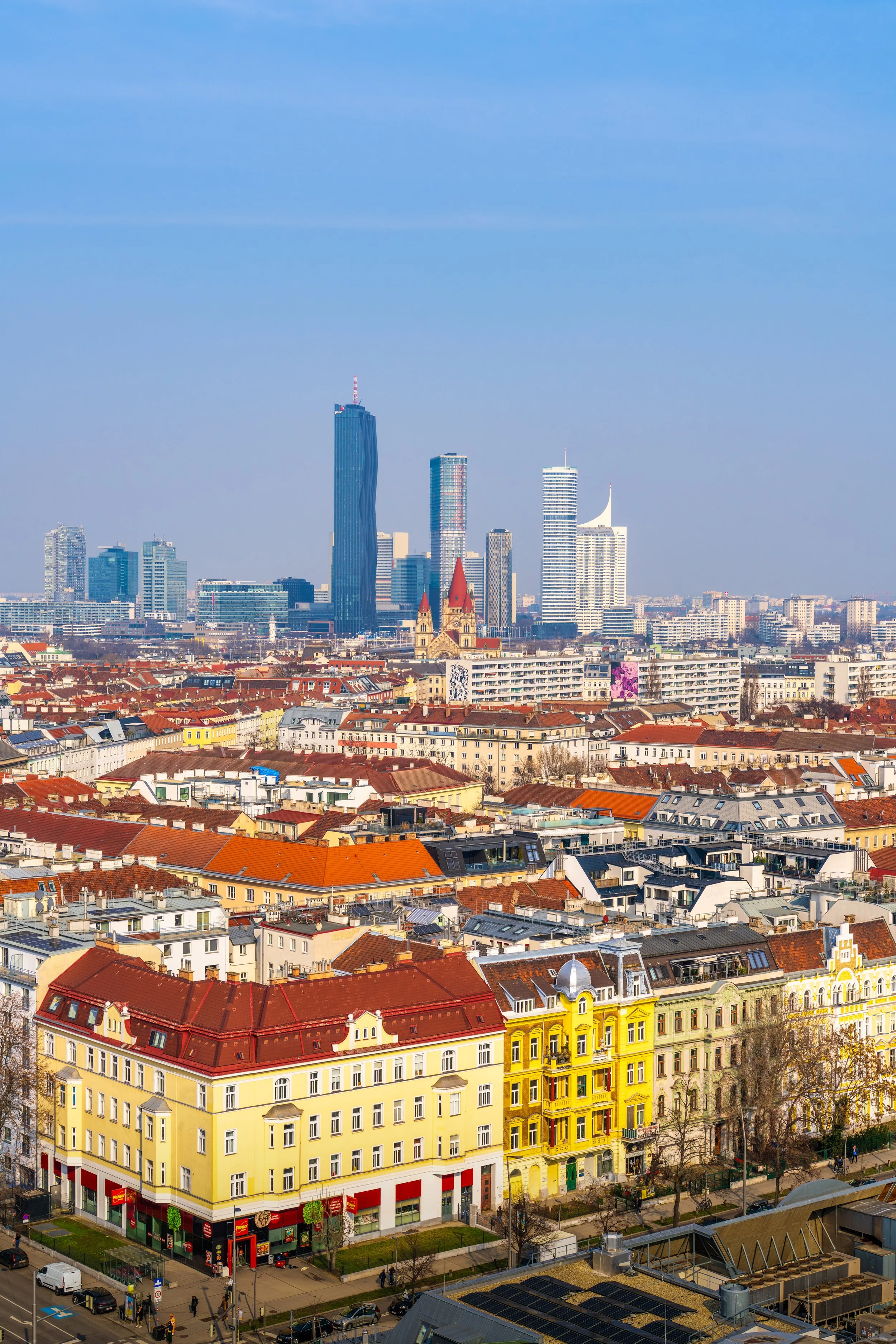 A detailed travel photography cityscape of Vienna, Austria, showcasing colorful historic buildings with red rooftops in the foreground and the modern skyline of Donau City in the background. The image captures the contrast between traditional Europea