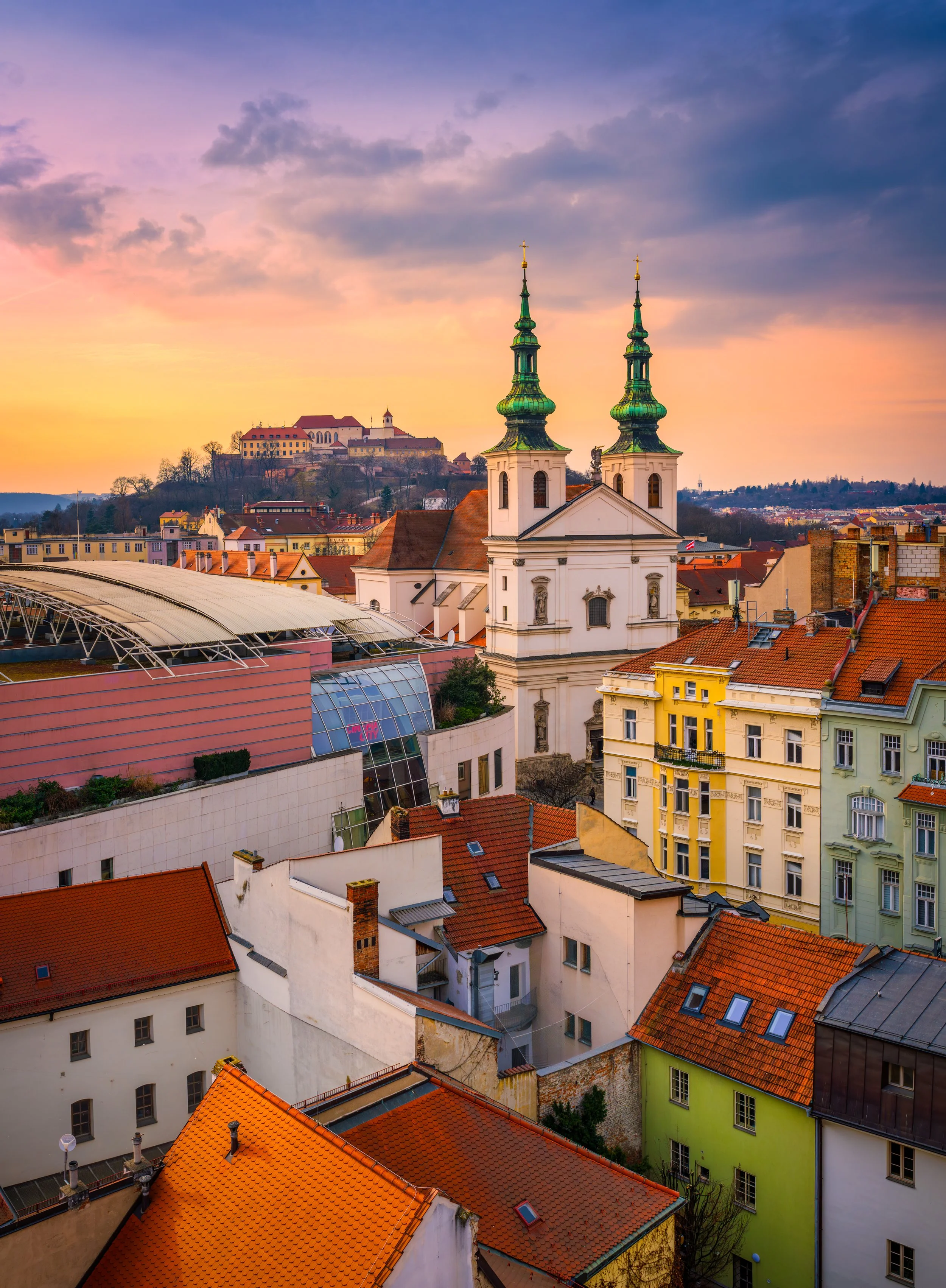 A vibrant travel photography image of Brno, Czech Republic, captured at sunset, featuring colorful rooftops, historic European architecture, and a prominent Baroque church with twin green towers. In the background, the iconic Špilberk Castle overlook