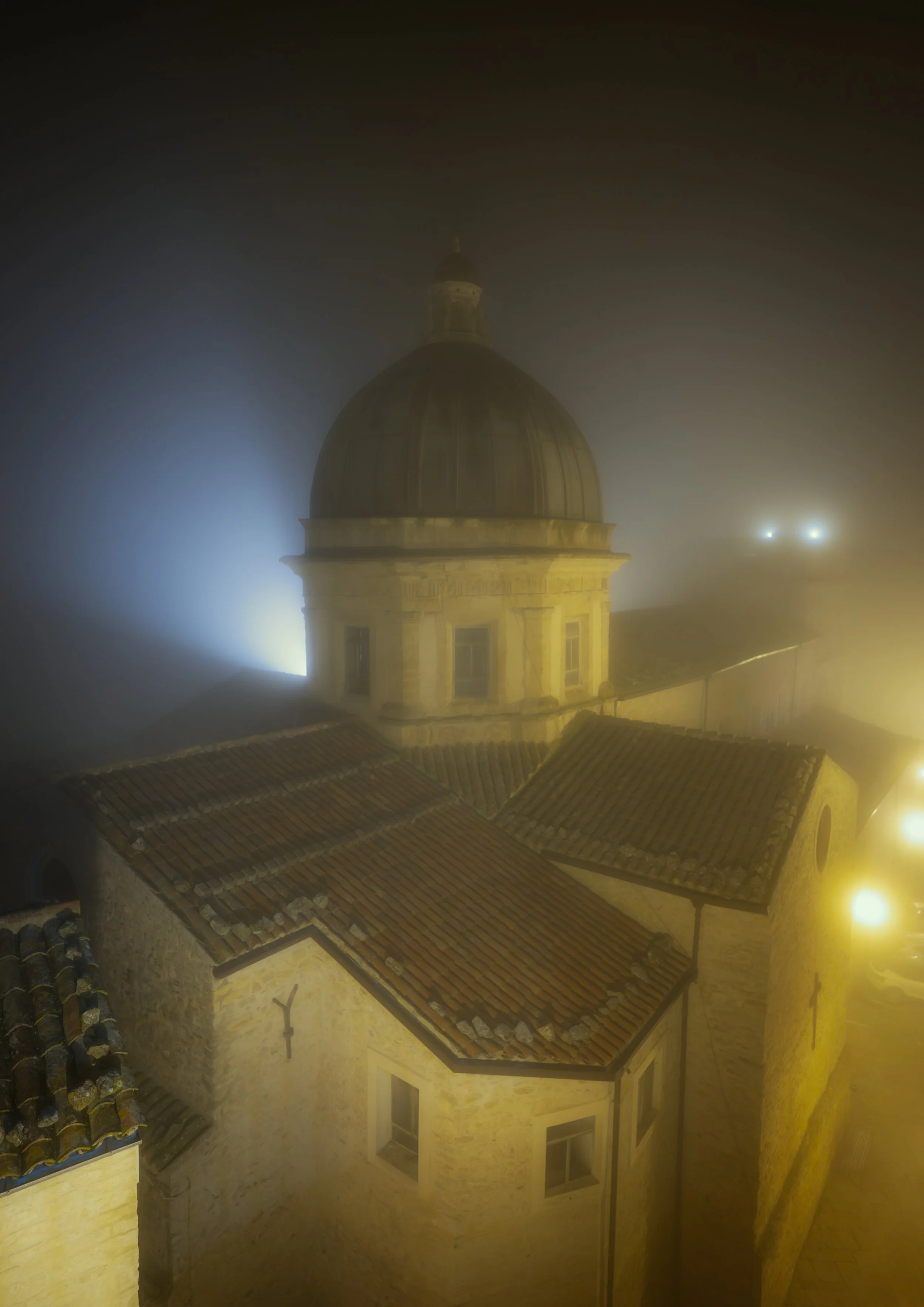 A foggy night scene of the historic church dome in Gangi, Sicily, softly illuminated by streetlights and diffused blue light. The mist enhances the atmosphere, revealing the stone walls, terracotta rooftops, and timeless Sicilian architecture. A capt