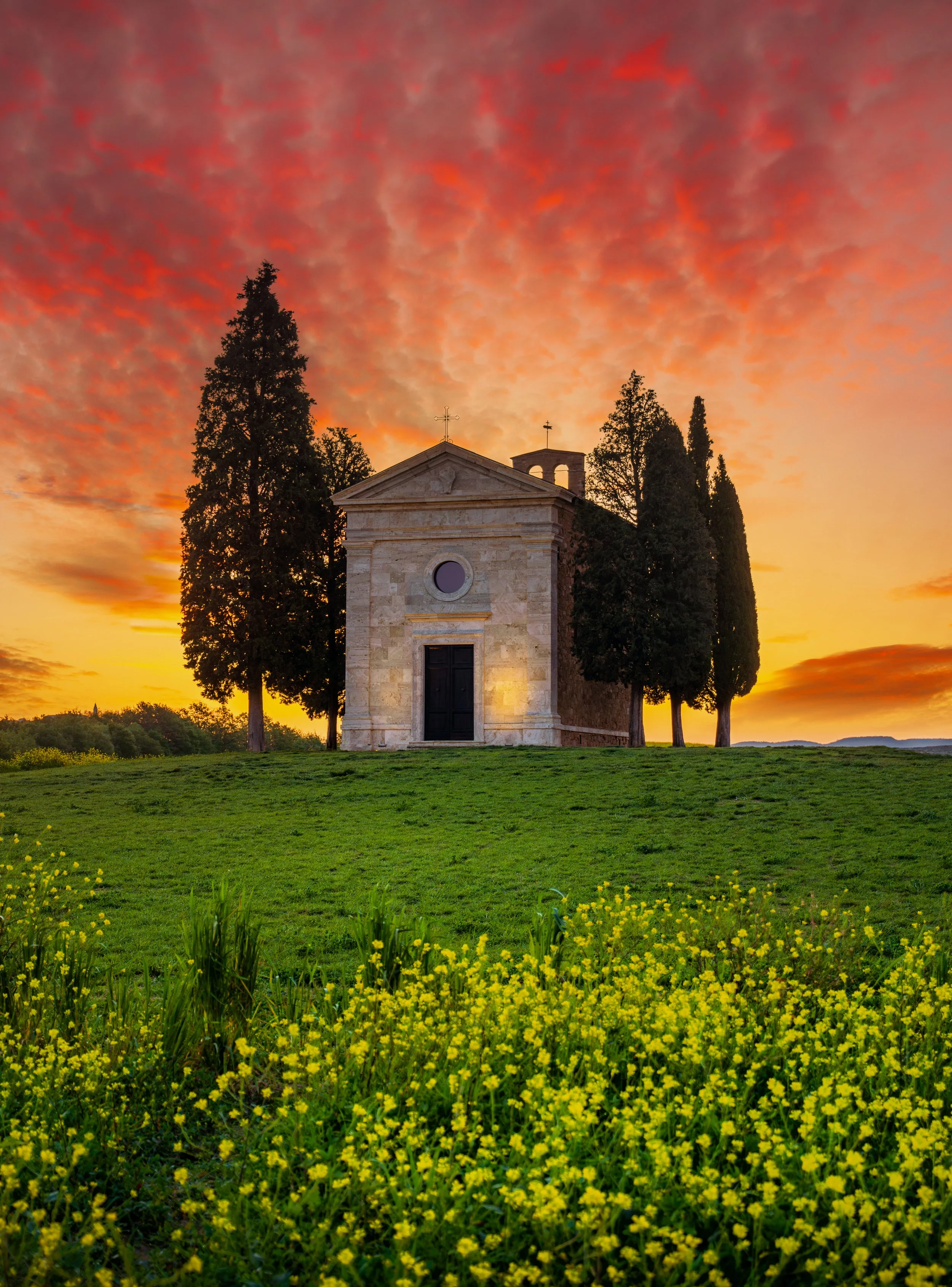 Sunrise over Chapel Vitaleta in Tuscany, surrounded by iconic cypress trees and blooming wildflowers. Dramatic sky, rich colors, and pure countryside calm —-Tuscany at its finest.