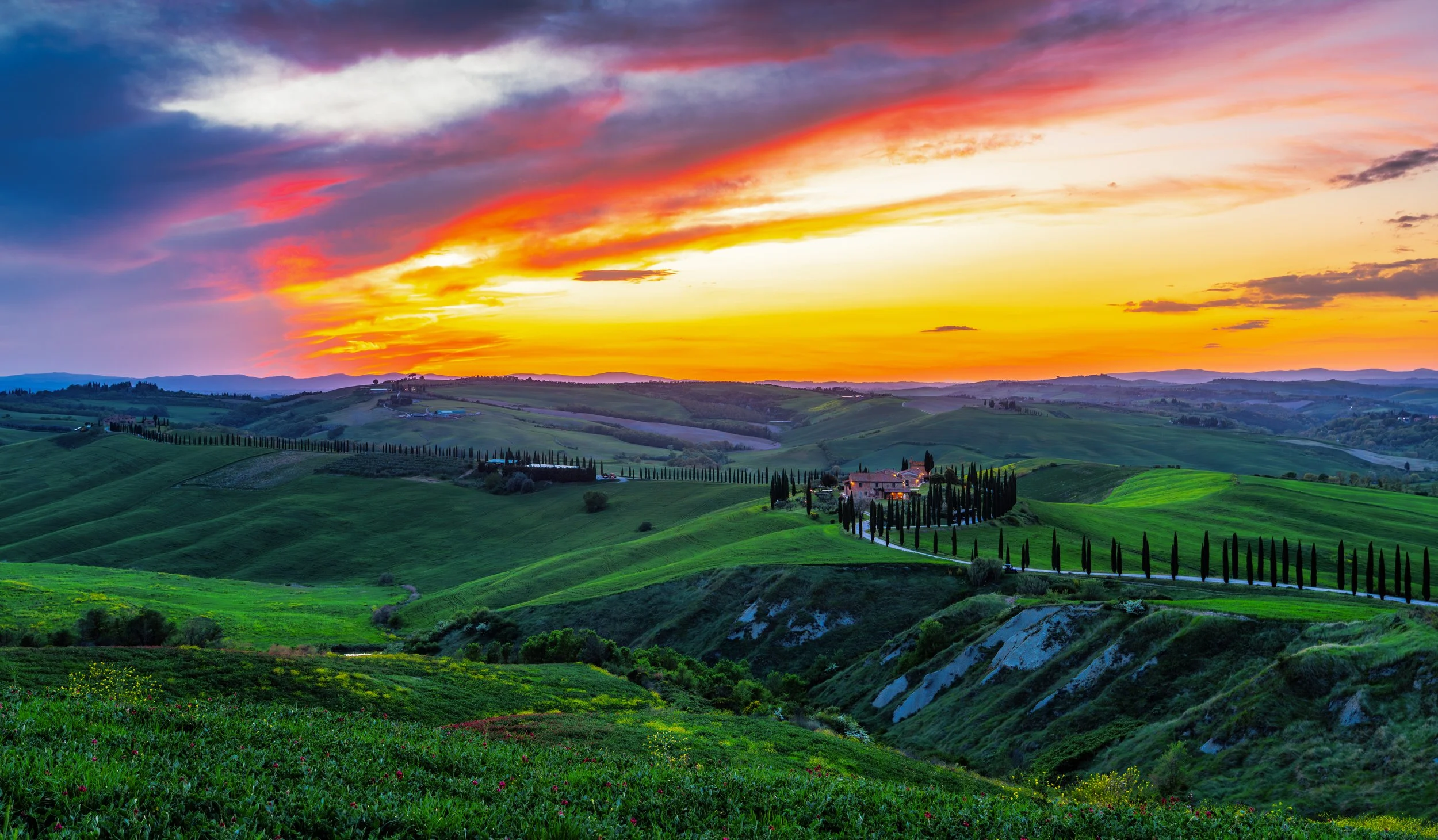 Breathtaking sunset over Val d’Orcia in Tuscany, featuring rolling green hills, a traditional farmhouse, and a winding road lined with cypress trees. The dramatic sky and warm light create one of the most iconic landscapes in Italy, perfect for trave