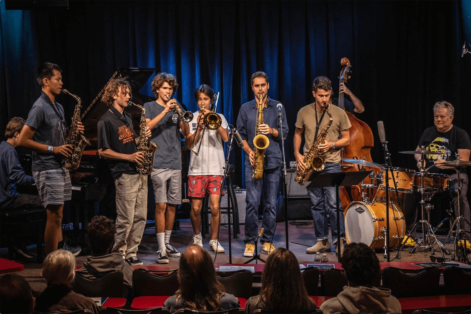 Ben Flocks performs as part of a jazz band on stage with eight other musicians playing various instruments including saxophones, trumpet, drums, double bass, and keyboard, with an audience watching.