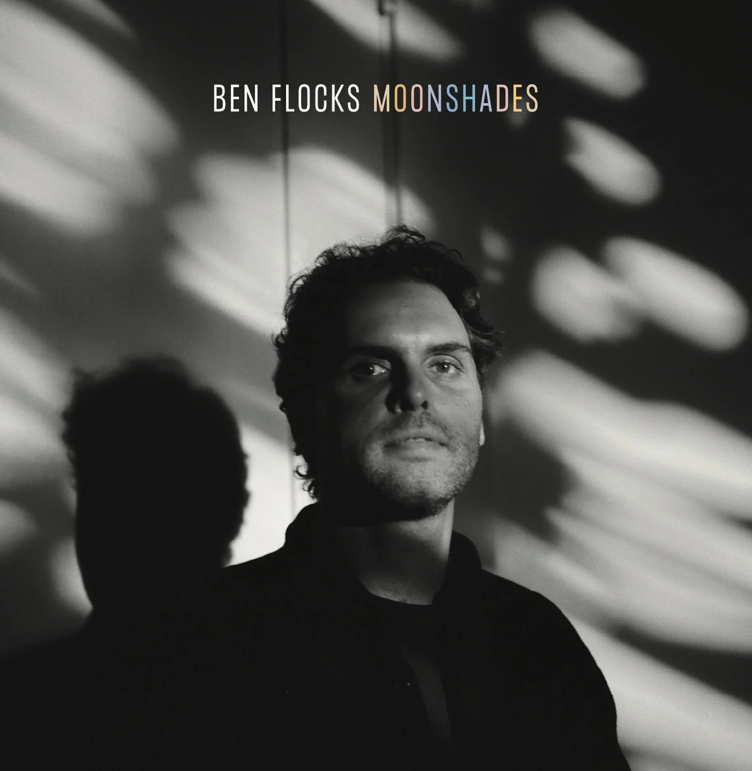 Black and white photo of Ben with serious expression, with shadows cast on the wall behind him. Text on the image reads 'Ben Flocks: Moonshades'.