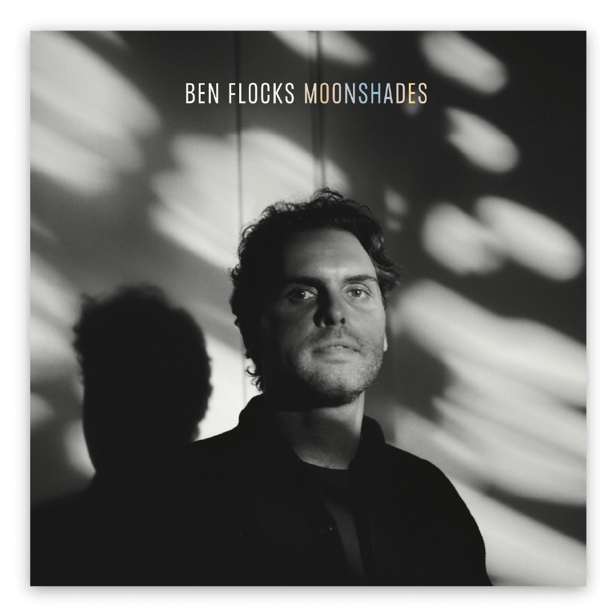 Black and white photo of Ben with serious expression, with shadows cast on the wall behind him. Text on the image reads 'Ben Flocks: Moonshades'.